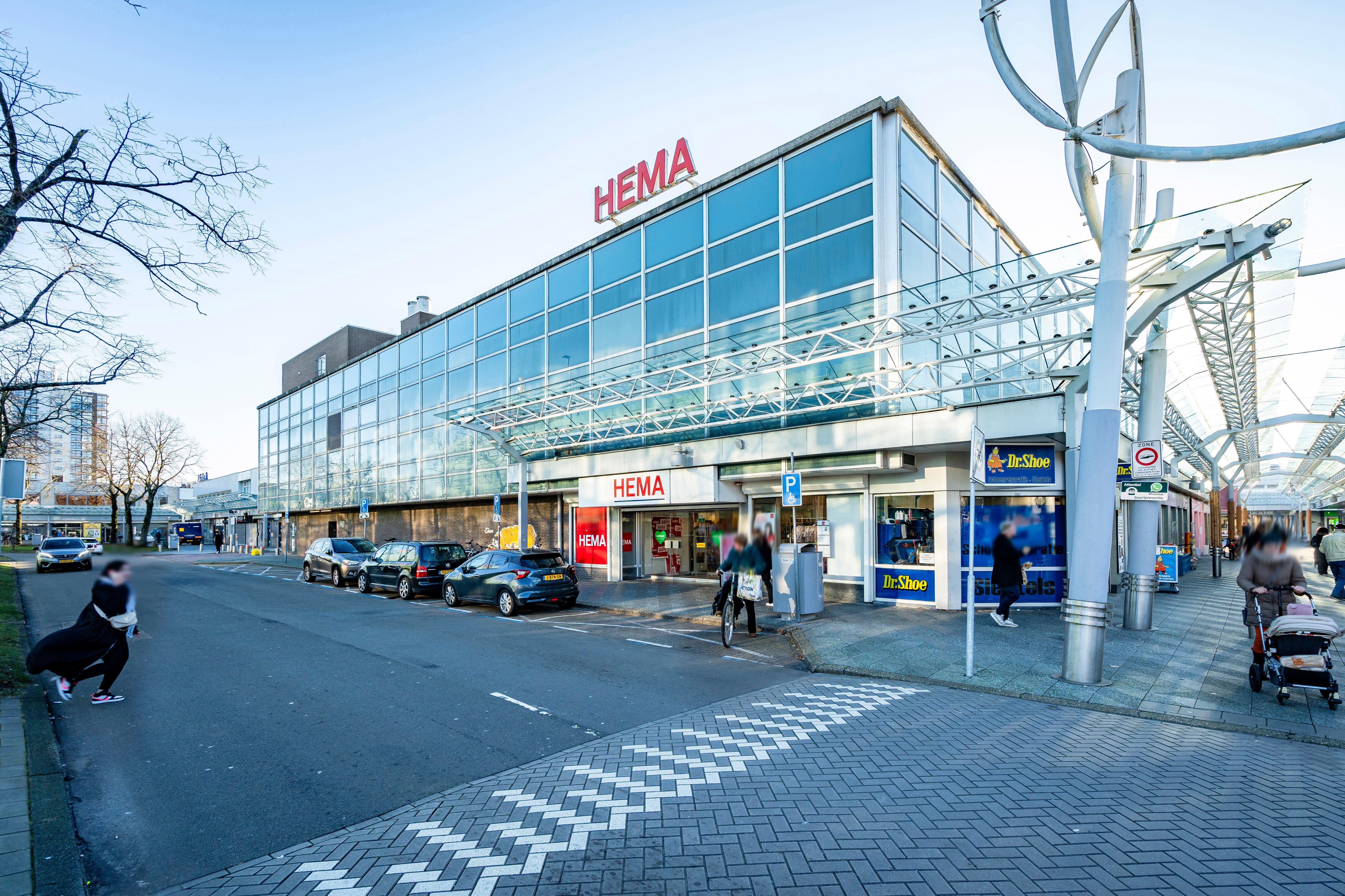 Een HEMA winkelpand met geparkeerde auto's en mensen die af en aan lopen en fietsen.