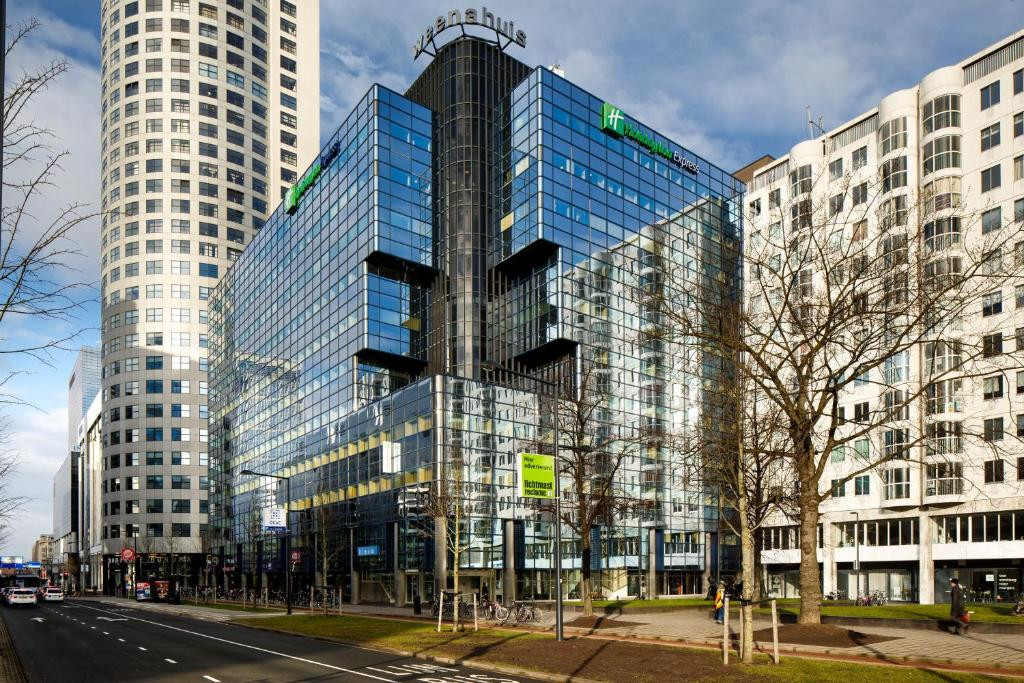 Een stadsgezicht met moderne gebouwen met glazen gevels onder een heldere lucht, inclusief een prominent gebouw van het “Holiday Inn” hotel, op een lege stedelijke straat.