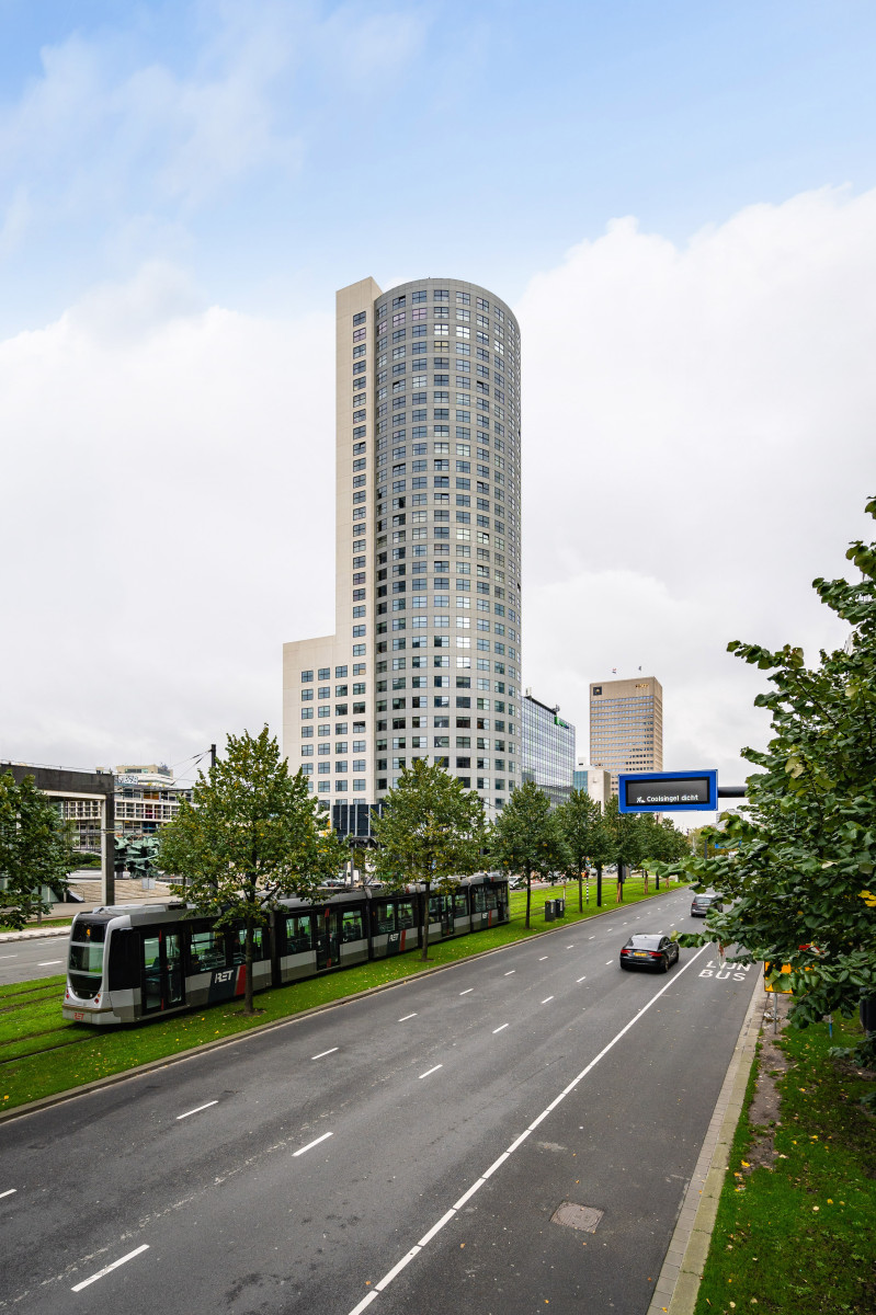 Een stedelijk tafereel met een hoge toren die prominent in het midden staat. De toren heeft een groot aantal ramen en lijkt voornamelijk uit woon- of kantoorruimtes te bestaan. Het is een modern gebouw met een lichtgekleurde gevel.