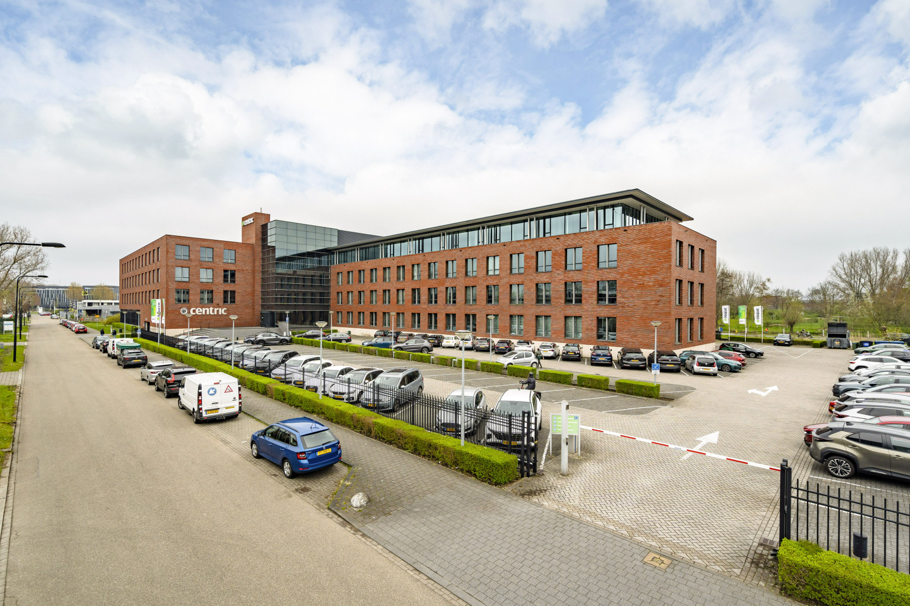 A modern office exterior located in a place outside of a city. The building has multiple levels large glas windows and a big parking lot with lots of cars parked.