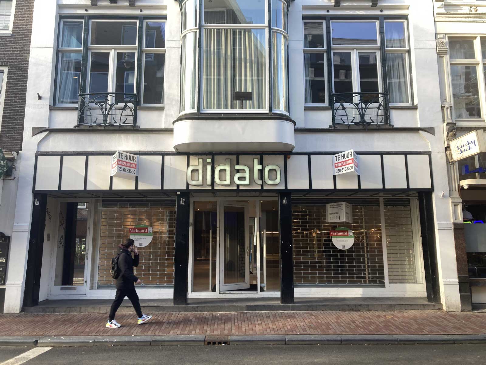 Een winkelpand heeft een drie verdiepingen tellende structuur met een witte gevel. Op de begane grond bevindt zich een grote glazen etalage en een glazen deur in het midden, beide omlijst met zwarte randen. Boven de deur en ramen zijn twee borden te zien met: 'TE HUUR'.  De eerste verdieping heeft twee symmetrische ramen met zwarte leuningen ervoor, en de tweede verdieping heeft een vergelijkbaar ontwerp maar zonder de leuningen. De ramen op zowel de eerste als de tweede verdieping hebben witte kozijnen en zijn onderverdeeld in kleinere vakken.  Het gebouw vertoont enkele Art Deco-invloeden, vooral merkbaar in de ronde erker die zich uitstrekt van de eerste naar de tweede verdieping boven de ingang. Deze erker heeft verticale roeden en een gebogen bovenkant die een elegante touch toevoegt aan de architectuur van het gebouw.  Voor het gebouw is een voetgangersstoep te zien. Een persoon loopt voorbij de etalage, vastgelegd in volle pas. De straat zelf oogt rustig zonder zichtbaar verkeer, en de lucht lijkt helder, wat suggereert dat het een zonnige dag zou kunnen zijn. De algehele indruk is van een rustige stedelijke omgeving.