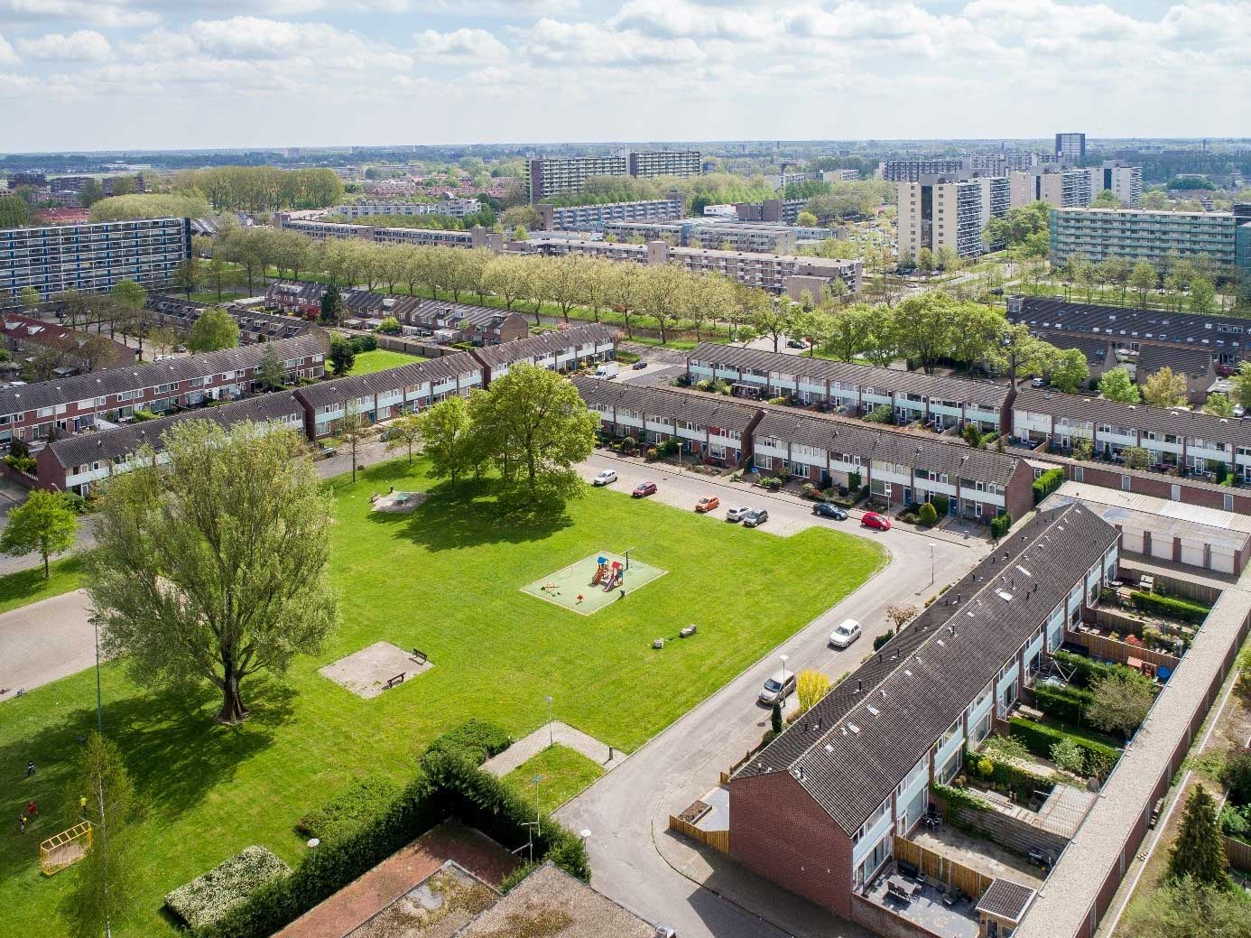 Luchtfoto van een buitenwijk met een centraal groen park, rijen huizen met rode daken, geparkeerde auto’s op straat en hoogbouwgebouwen op de achtergrond onder een heldere hemel