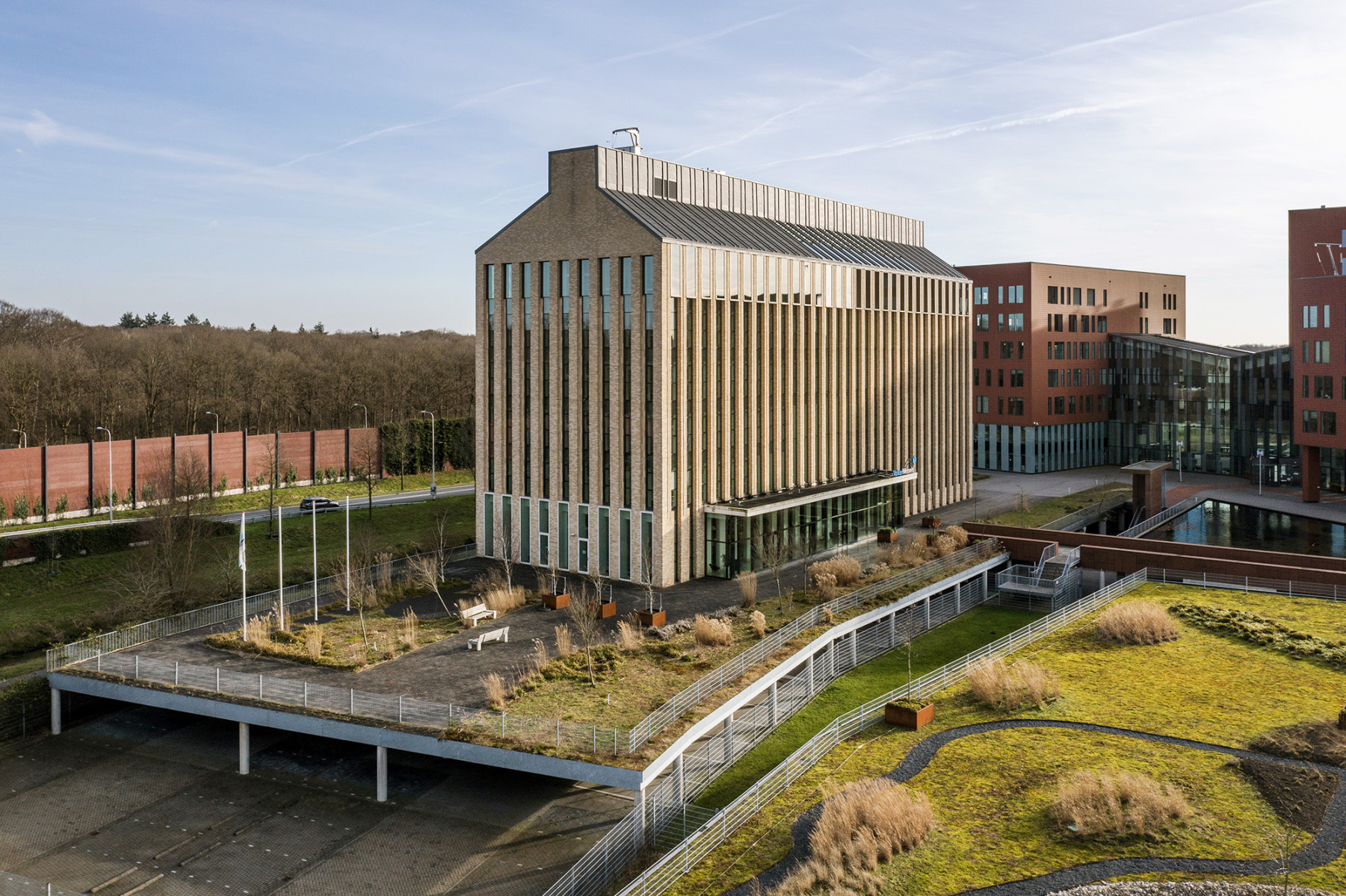 Een modern gebouw vanuit een verhoogd perspectief. Het gebouw lijkt meerdere verdiepingen te hebben en heeft een rechthoekige vorm met een uitgesproken geometrisch patroon op de gevel, bestaande uit verticale lijnen en rechthoekige vormen. De kleur van het gebouw is een mix van natuurlijke steentinten, variërend van licht tot donker.