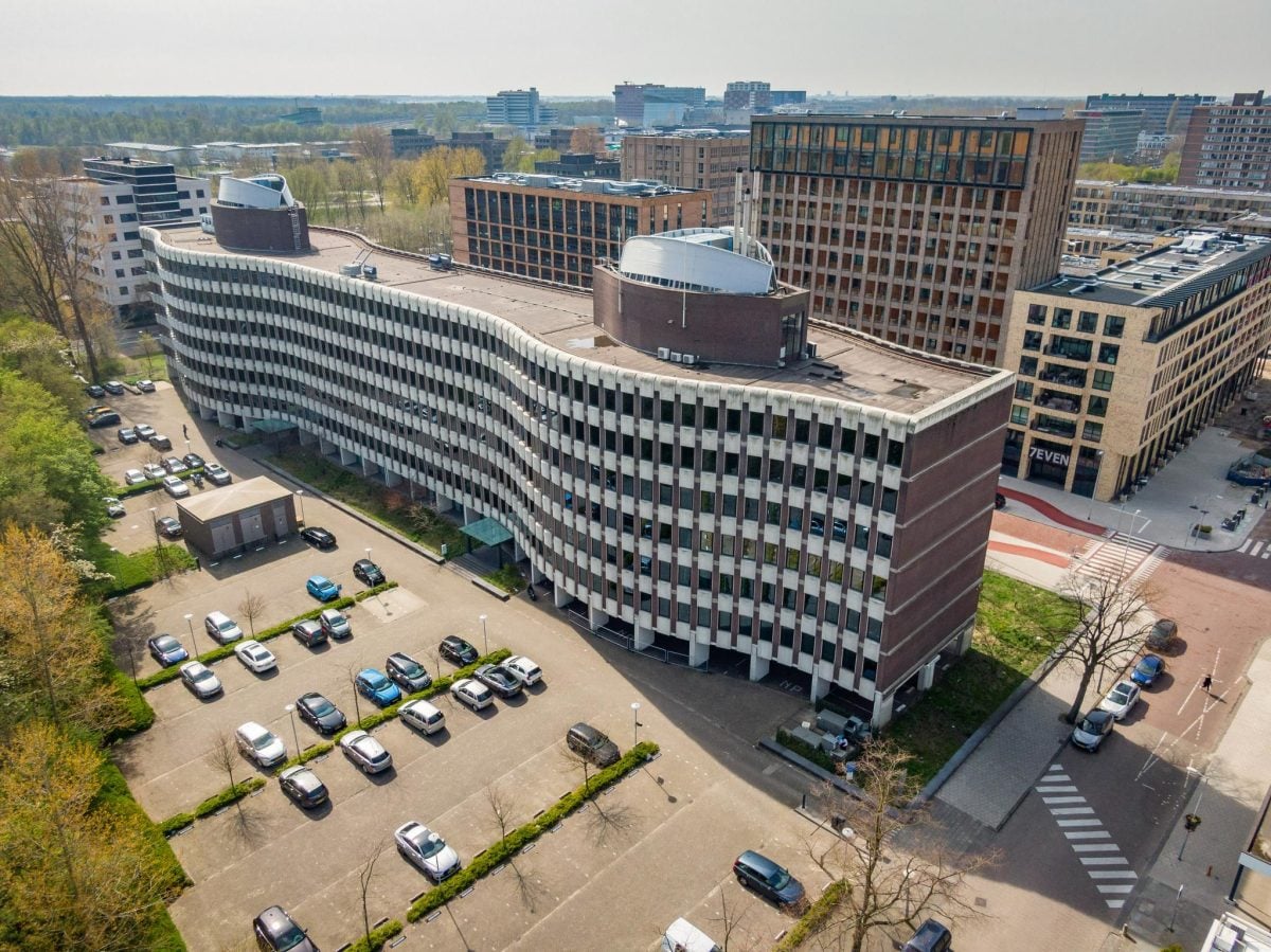 Luchtfoto van een modern stadslandschap met een uniek golvend ontworpen gebouw met witte gevel, omringd door andere structuren, parkeerplaatsen vol met auto’s, groen en een heldere lucht erboven. 