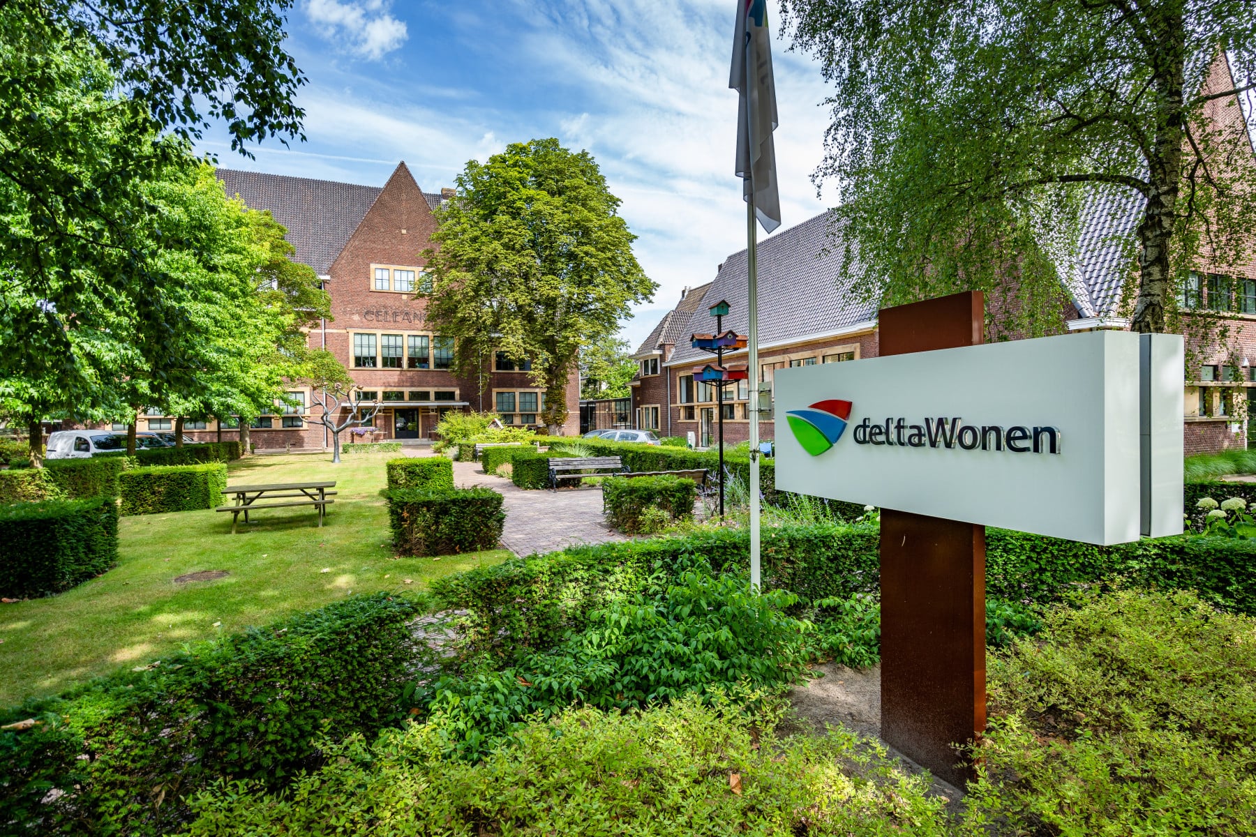 Een weelderige groene tuin voor het bakstenen gebouw van deltaWonen, met bankjes, verzorgde gazons en bomen, met op de voorgrond een ‘deltaWonen’-bord.