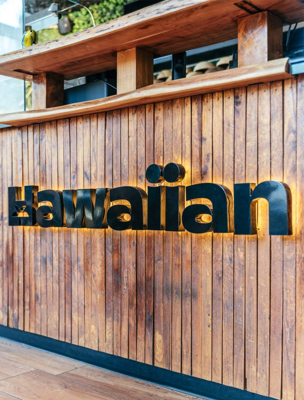 Een bar gemaakt van houten latten die verticaal geplaatst zijn. Op de bar staat de naam 'Hawaiian' met zwarte letters en omgeven door geel licht.