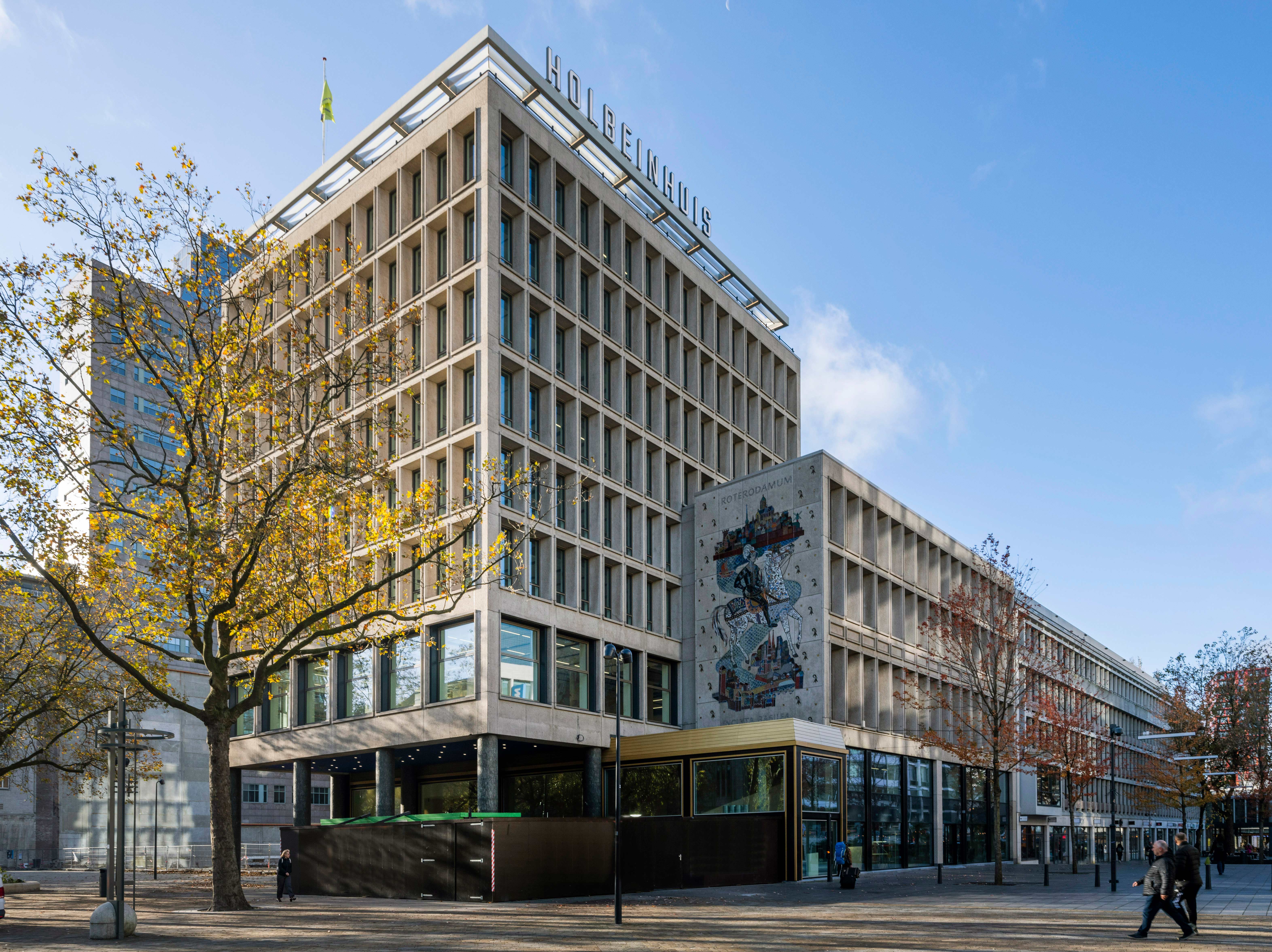 stadsgezicht met een prominent gebouw, een café of winkel aan de voorkant, een groot mozaïek of kunstwerk op de muur, bomen met herfstbladeren, twee personen op de stoep, en een heldere, zonnige dag met een blauwe lucht. De setting straalt een levendige stedelijke sfeer uit met moderne architectuur en artistieke elementen.