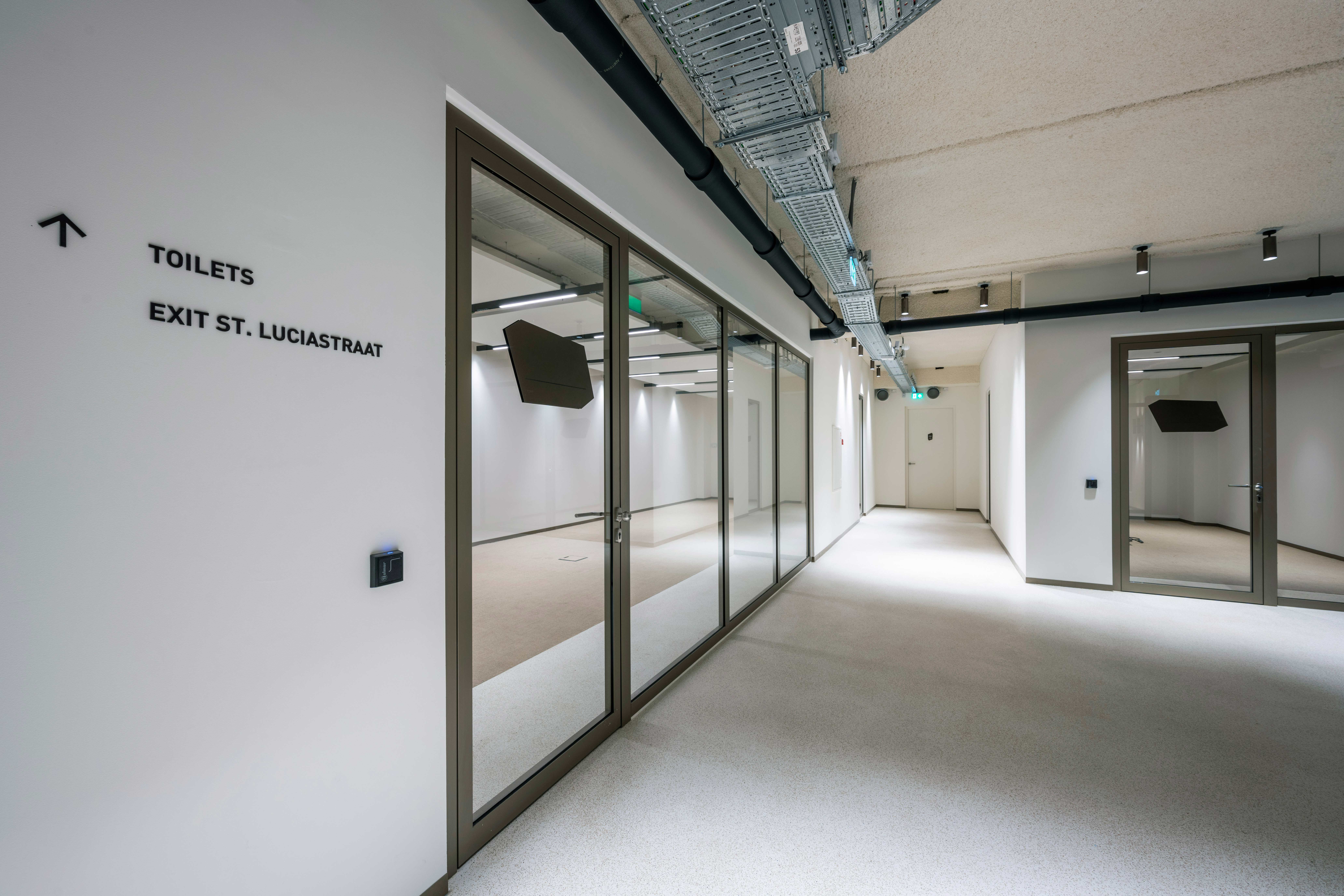 Een goed onderhouden commerciële gang met lichte muren, een blootgesteld betonnen plafond met hangende installaties, een licht tapijt op de vloer, inbouwspots voor verlichting, bewegwijzering naar toiletten en een uitgang, glazen deuren en ramen met zwarte frames, en een eigentijdse en professionele uitstraling.