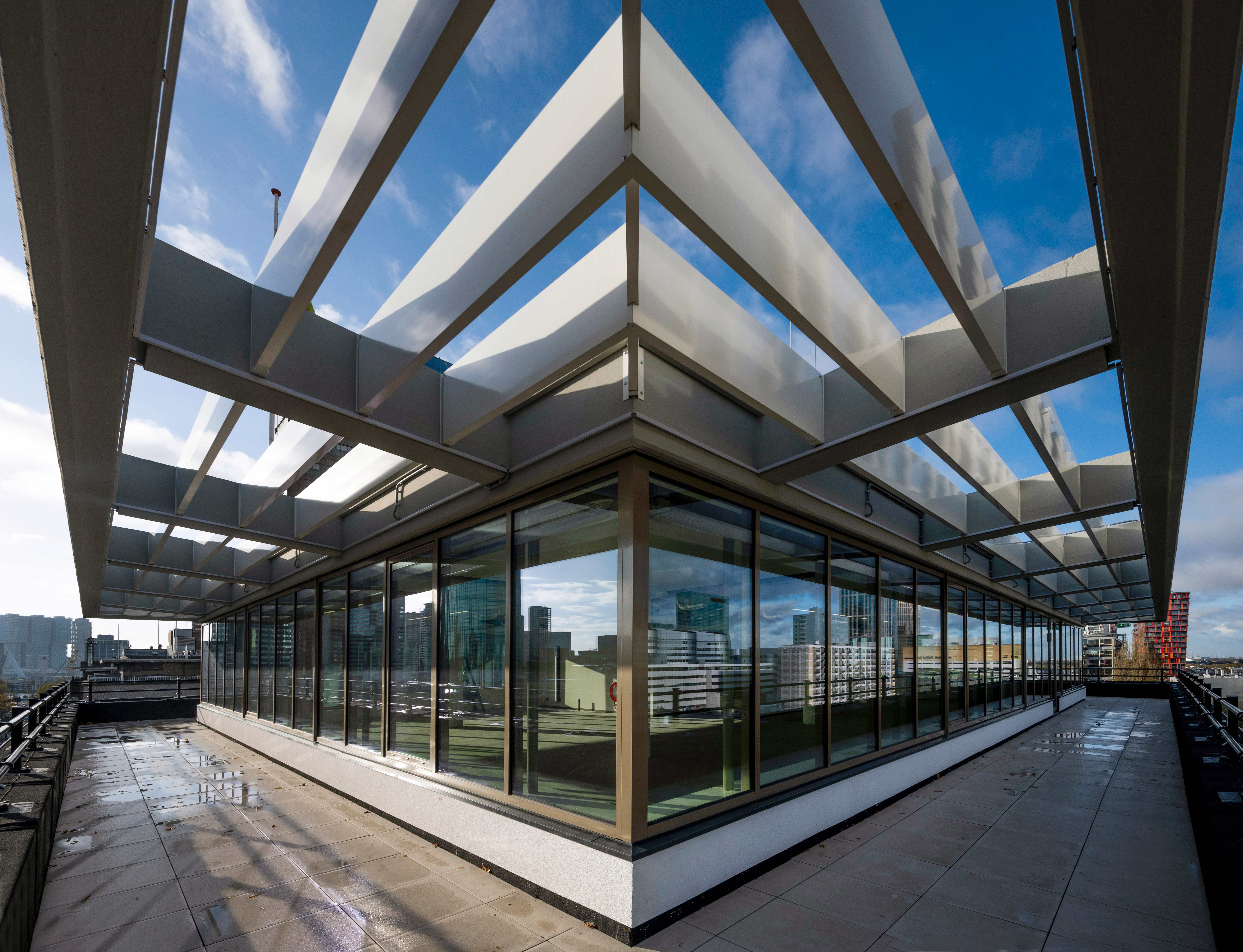 Een moderne glazen omheining met een geometrisch ontwerp, ondersteund door witte balken en diagonale steunberen op een terras of dak met uitzicht op een stedelijke omgeving. De omheining reflecteert de omliggende gebouwen en de lucht, terwijl de steunberen schaduwen werpen op het glas eronder. De omgeving straalt een gevoel van moderniteit, transparantie en architectonische elegantie uit.