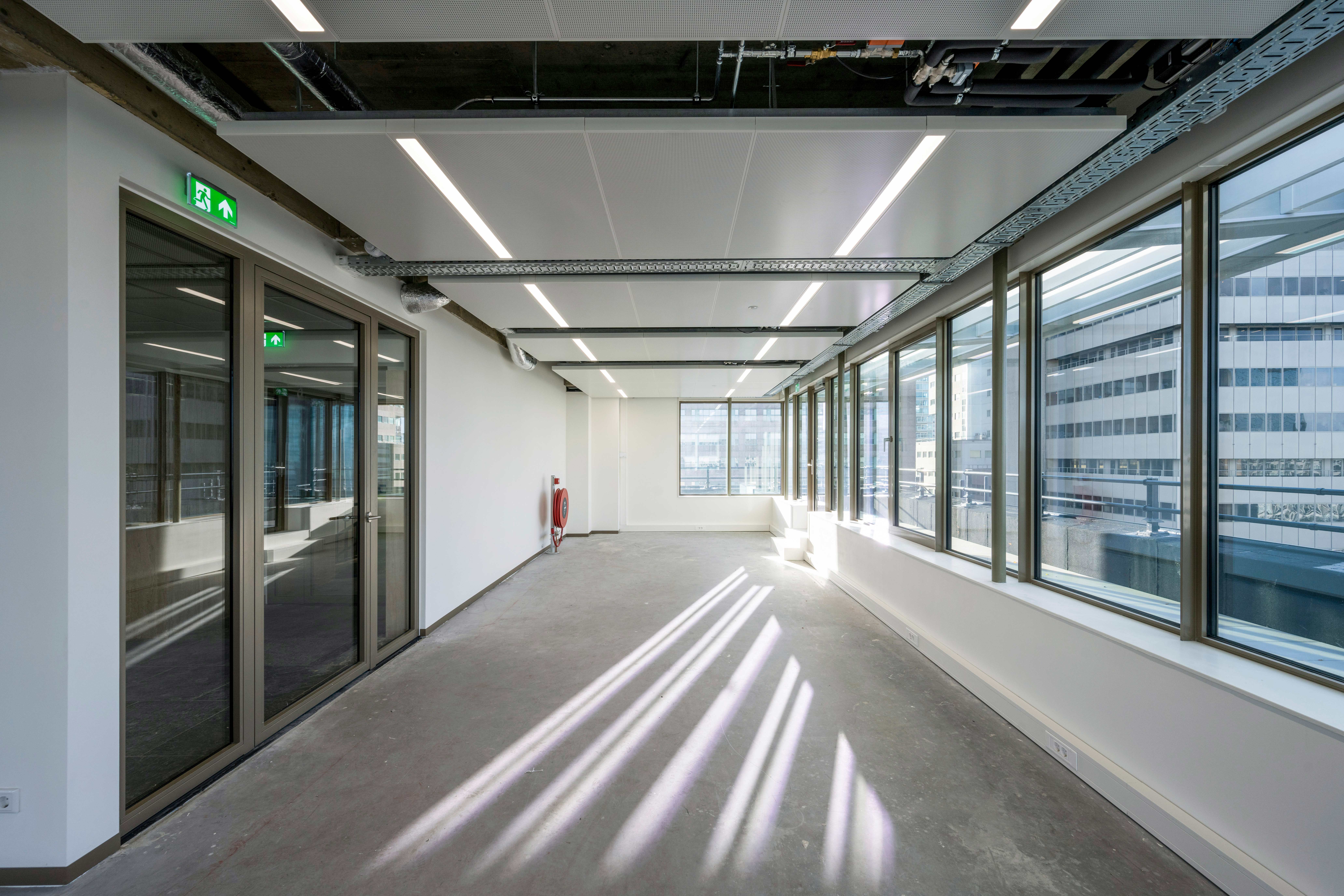 Een leeg kantoor of commerciële ruimte met grote ramen aan de rechterkant die uitzicht bieden op nabijgelegen gebouwen. De ruimte heeft een witte betonnen vloer, witte muren, en een blootgelegd plafond met industriële elementen. Een rood brandblusapparaat en een groene nooduitgangdeur zijn aanwezig. De ruimte is minimalistisch en klaar voor gebruik.