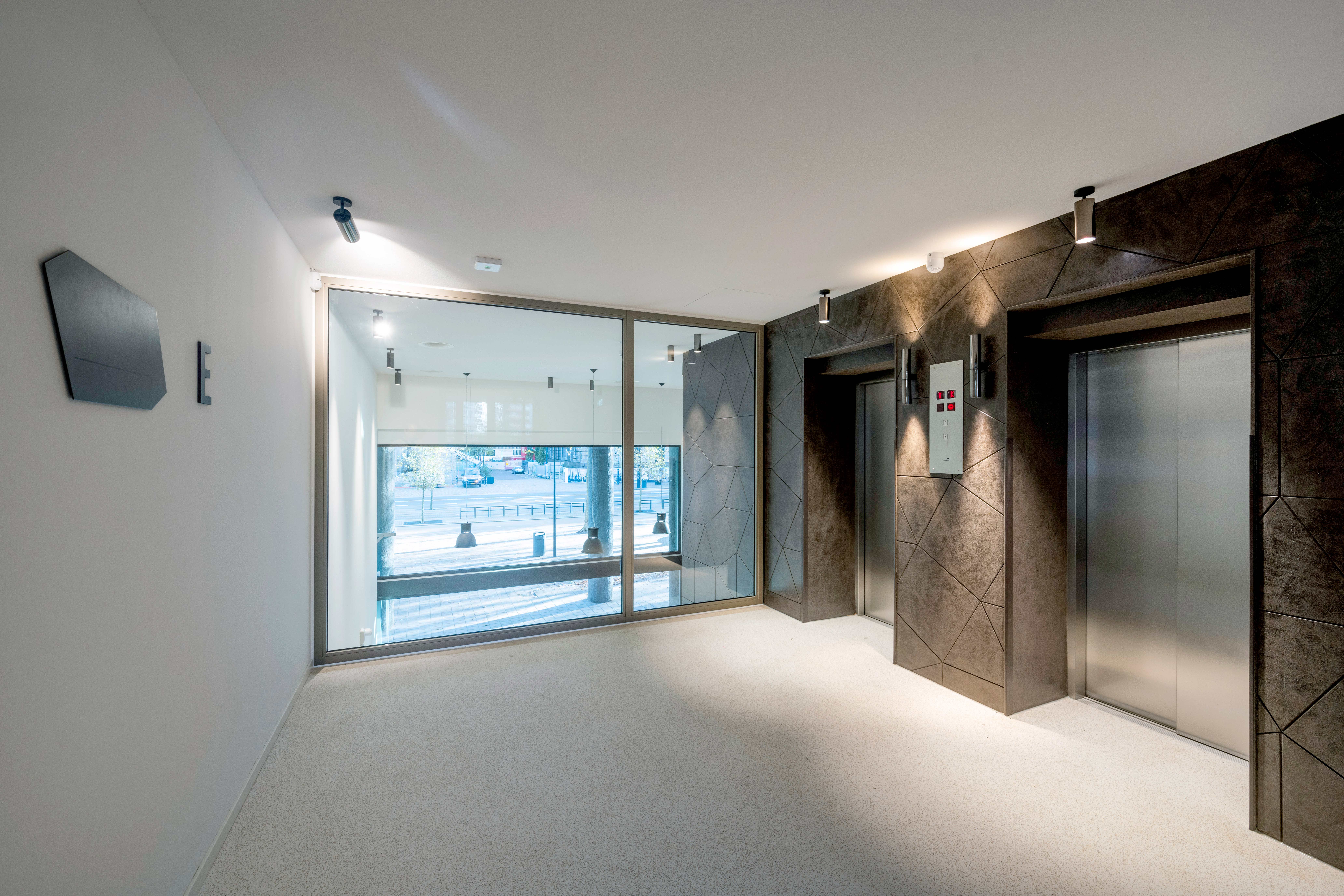 Een liftlobby met een lichtgekleurd, gespikkeld tapijt op de vloer. Aan de linkerkant is een wandgemonteerde flatscreen display te zien en aan de rechterkant bevinden zich twee liftdeuren van geborsteld metaal, omlijst met donker marmer. Tussen de liften is een liftoproeppaneel met knoppen. De lobby heeft een glazen achterwand die natuurlijk licht binnenlaat, en er zijn inbouw plafondlampen aanwezig. Het plafond is een wit systeemplafond. De sfeer is modern, minimalistisch en functioneel.