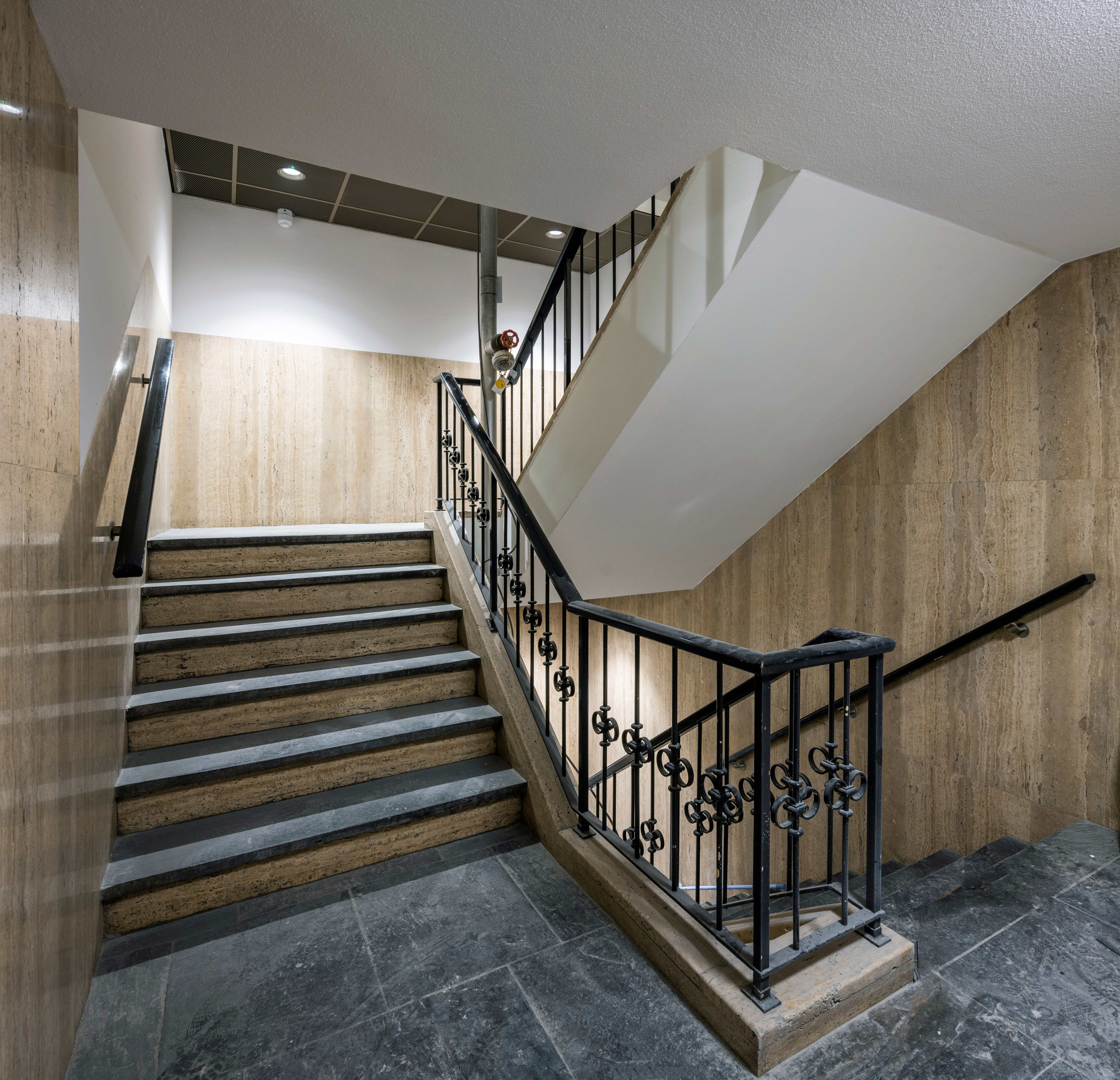 Een trap in een binnengebouw met beige wanden, zwarte leuningen met decoratieve elementen, en treden met een combinatie van donkere en lichte stenen.