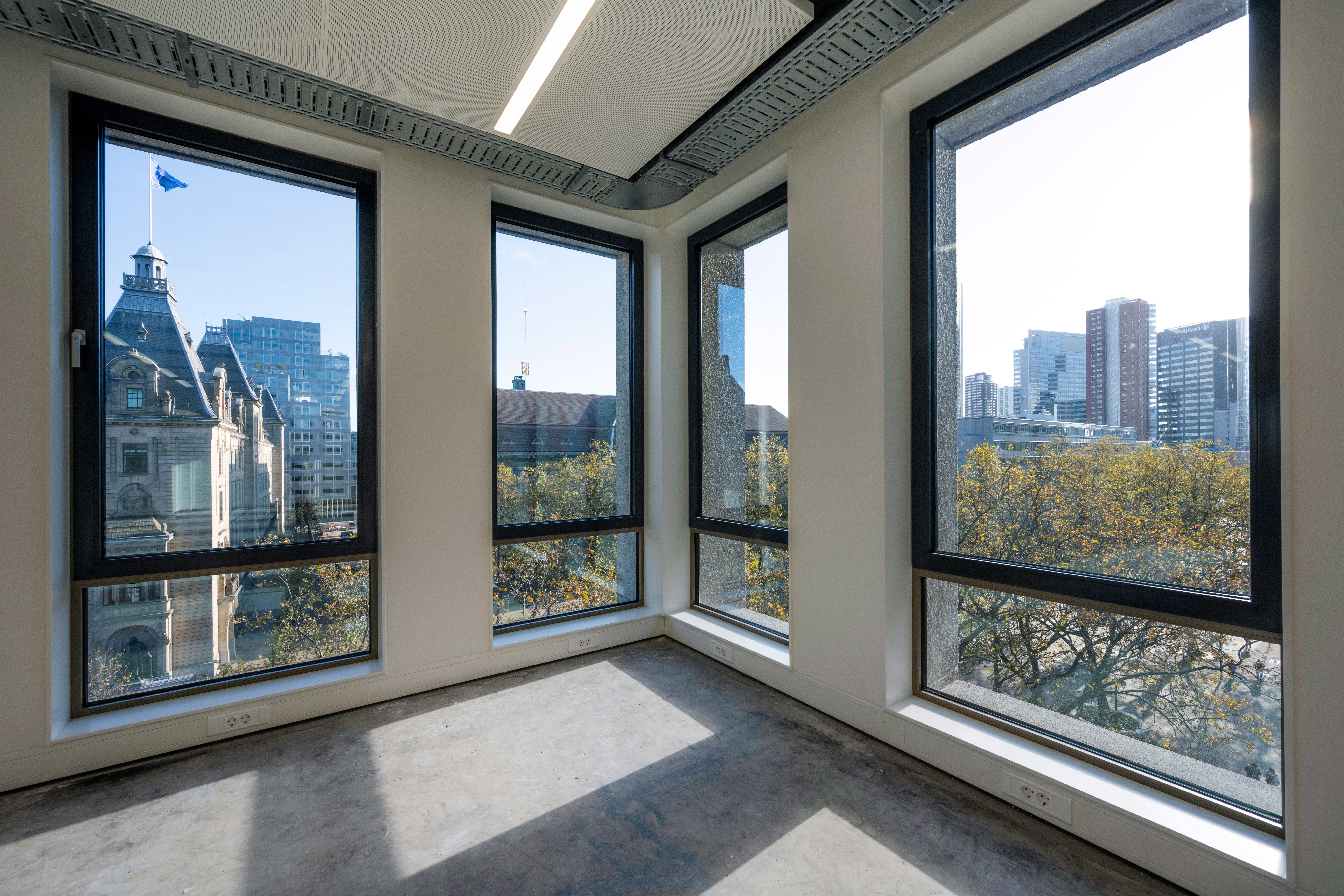 Een lege kamer in een hoog gebouw met grote ramen die uitzicht bieden op een stadslandschap met een historisch stenen gebouw aan de linkerkant en moderne gebouwen aan de rechterkant. De kamer heeft betonnen vloeren, witte muren, en een minimalistische inrichting, met veel natuurlijk licht dat de ruimte vult.