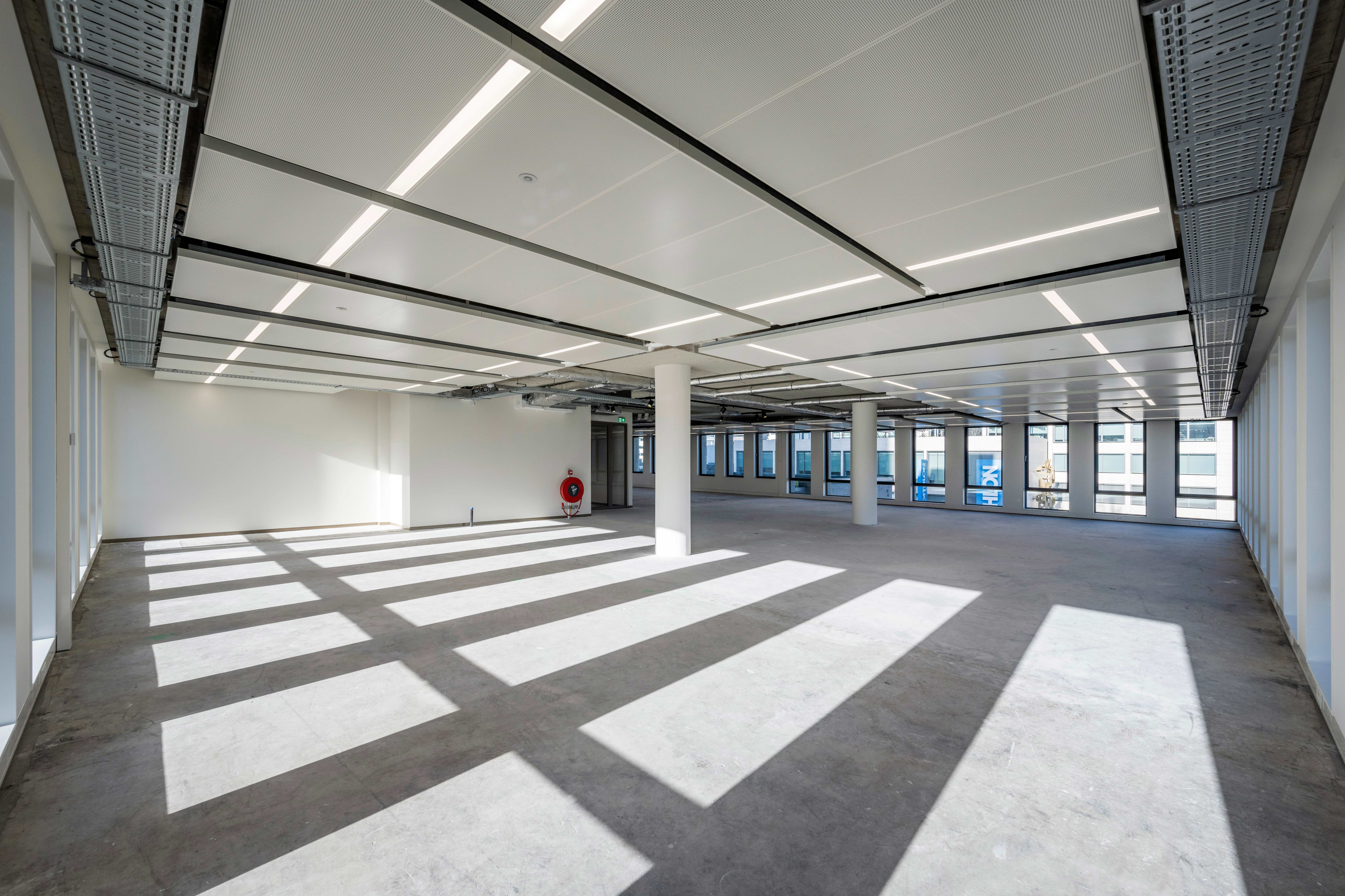 Een ruime, lege binnenruimte met een modern industrieel plafond met zichtbare leidingen en kabelgoten, gepolijste betonnen vloer, witte muren en grote ramen die natuurlijk licht binnenlaten. LED-panelen verlichten de ruimte en werpen scherpe schaduwen van de raamkozijnen op de vloer. Een rode brandblusser is aan de rechterwand gemonteerd, en witte kolommen verspreid over de ruimte. De ruimte straalt een schone, open uitstraling uit, klaar voor gebruik of aanpassing aan de behoeften van een bedrijf of huurder.