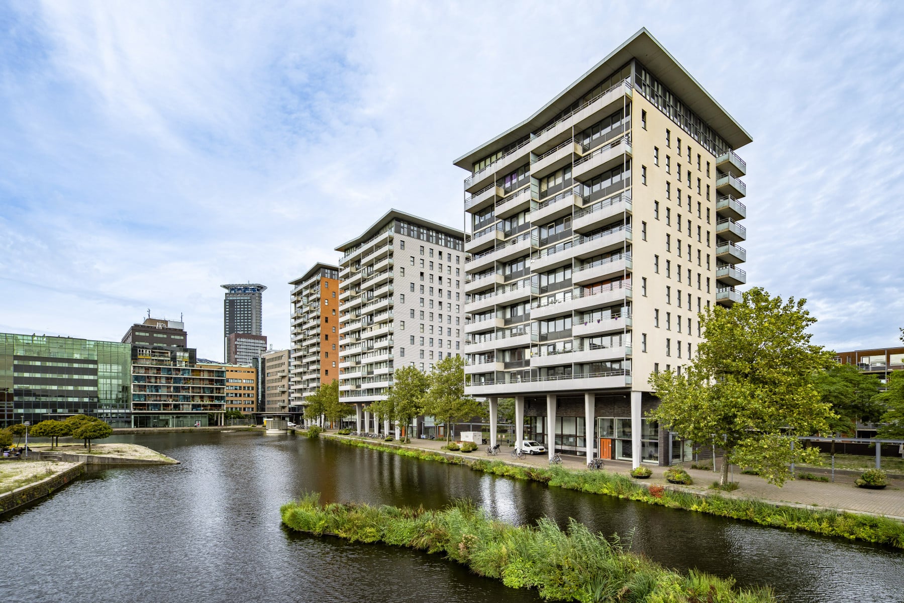Een stedelijke waterkant met een reeks van hoge gebouwen die variëren in hoogte en ontwerp. Deze gebouwen zijn voornamelijk residentiële appartementencomplexen, met een paar kantoorpanden aan de linkerkant van het beeld. De architectuur is hedendaags, met een mix van balkons, ramen en verschillende gevelmaterialen.  De gebouwen staan langs de rand van een kanaal of rivier, met kalme waterspiegeling die de structuren gedeeltelijk weerspiegelt. Aan de overkant van het water is er een goed onderhouden groenstrook met gras, struiken en enkele kleine bomen. Het lijkt een aangelegd gebied te zijn, mogelijk bedoeld als esthetische buffer tussen het water en de bebouwing.  De lucht is overwegend bewolkt, maar het is licht genoeg om te suggereren dat de foto overdag is genomen. Er zijn geen mensen zichtbaar in de scène, wat een rustige en misschien ietwat verlaten indruk geeft. De algehele sfeer lijkt vredig en geordend.