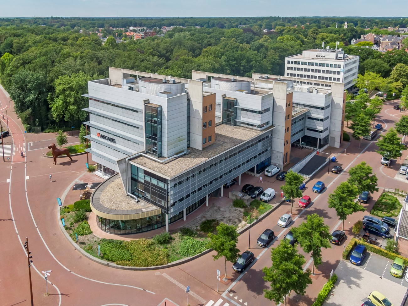Een modern gebouwencomplex met meerdere niveaus en secties. De architectuur omvat een mix van rechte en gebogen lijnen, met een opvallende gebogen glazen gevel aan één uiteinde. Het gebouw wordt omringd door groen, wegen en geparkeerde auto’s, wat aangeeft dat het zich in een stedelijk gebied bevindt. Er zijn geen zichtbare wiskundige of huiswerkproblemen in de afbeelding om te transcriberen.