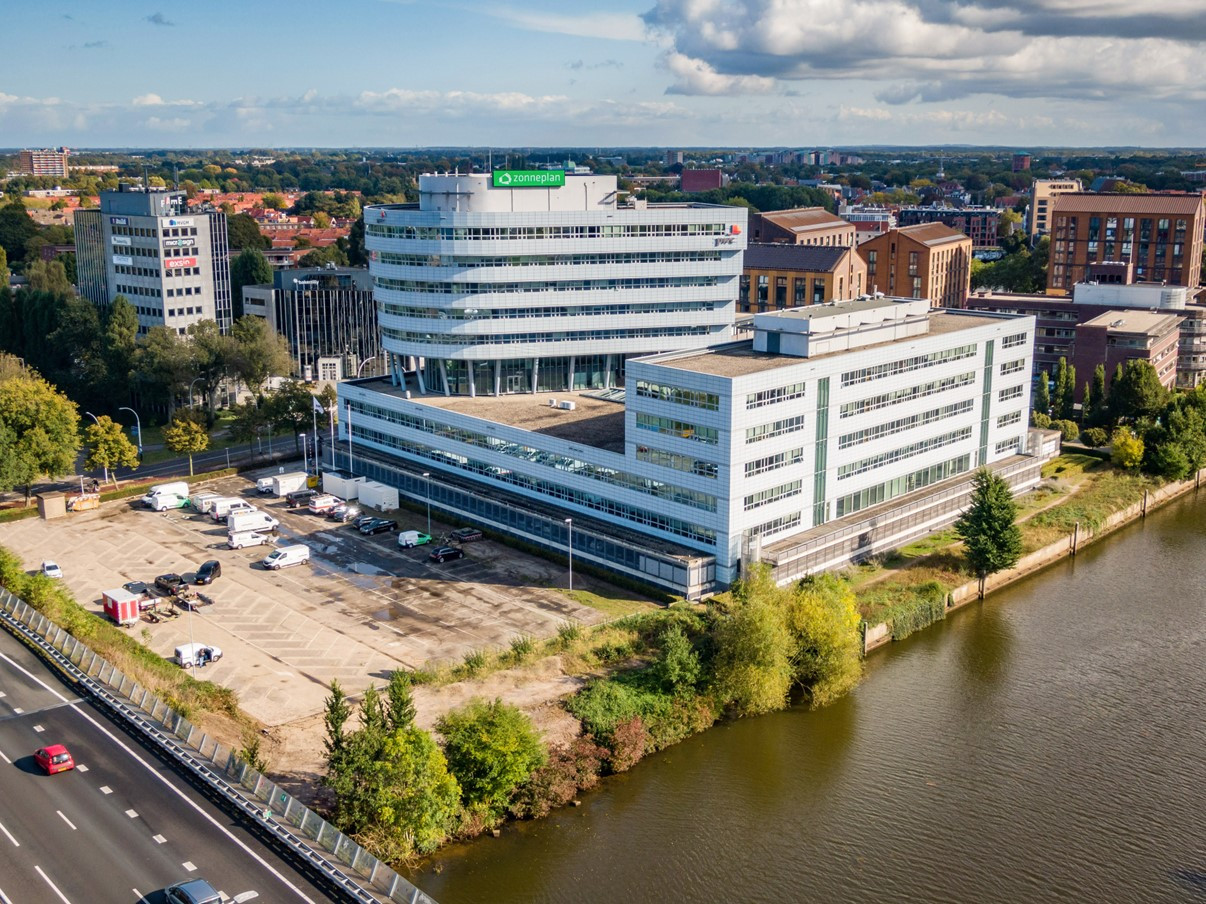 Een luchtfoto van een modern gebouw met meerdere verdiepingen naast een waterpartij, omringd door stedelijke bebouwing en groen.