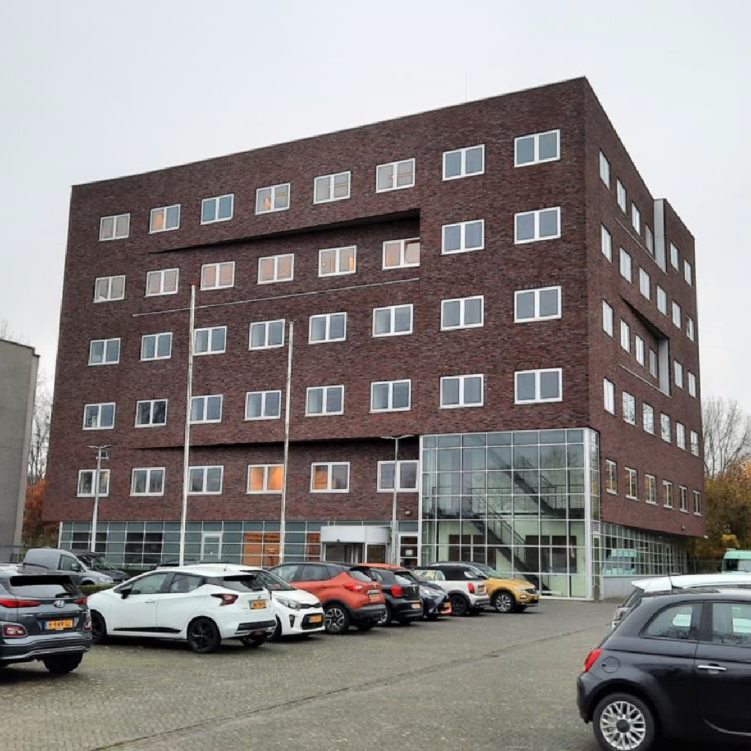 Een modern, meerdere verdiepingen tellend bakstenen gebouw met veel ramen, omgeven door een parkeerplaats en andere gebouwen.