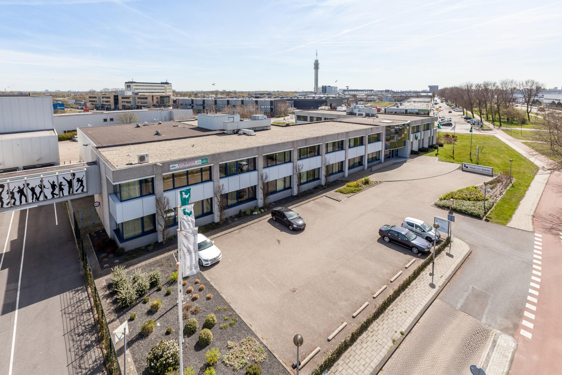 Een tweelaags gebouw met een plat dak. Het object staat aan een geasfalteerde weg met verschillende rijstroken en verkeersmarkeringen. Voor het gebouw is een parkeerplaats met een paar auto's. Aan de linkerkant van het gebouw is een rij silhouetten van mensen aangebracht, wat lijkt op een kunstwerk of decoratie. Het gebouw heeft grote ramen en de architectuur is functioneel en modern.