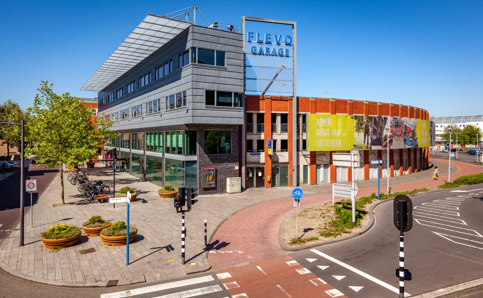 Een eigentijds meerdere verdiepingen tellend gebouw met de tekst 'FLEVO GARAGE' aan een stedelijke straatkruising onder een heldere blauwe lucht.