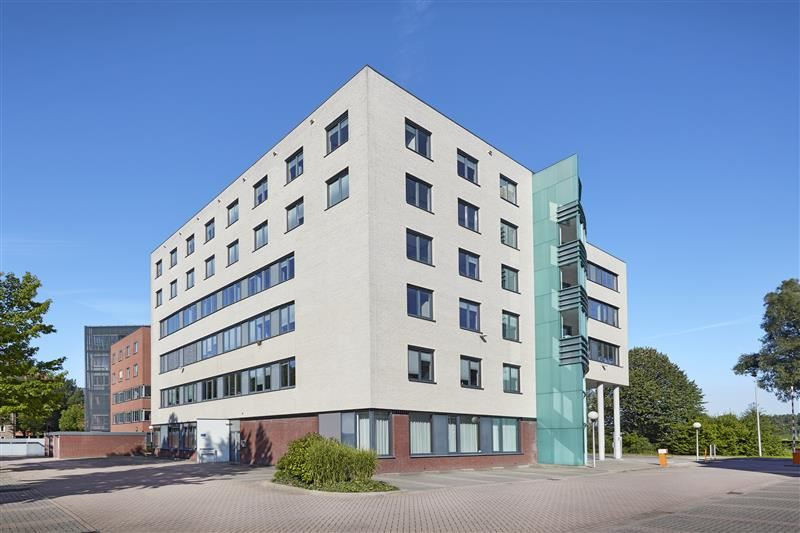 Een modern wit meerdere verdiepingen tellend gebouw onder een heldere blauwe lucht, met een externe groene trapstructuur op de gevel, een ander bakstenen gebouw op de achtergrond en een verharde voorgrond.