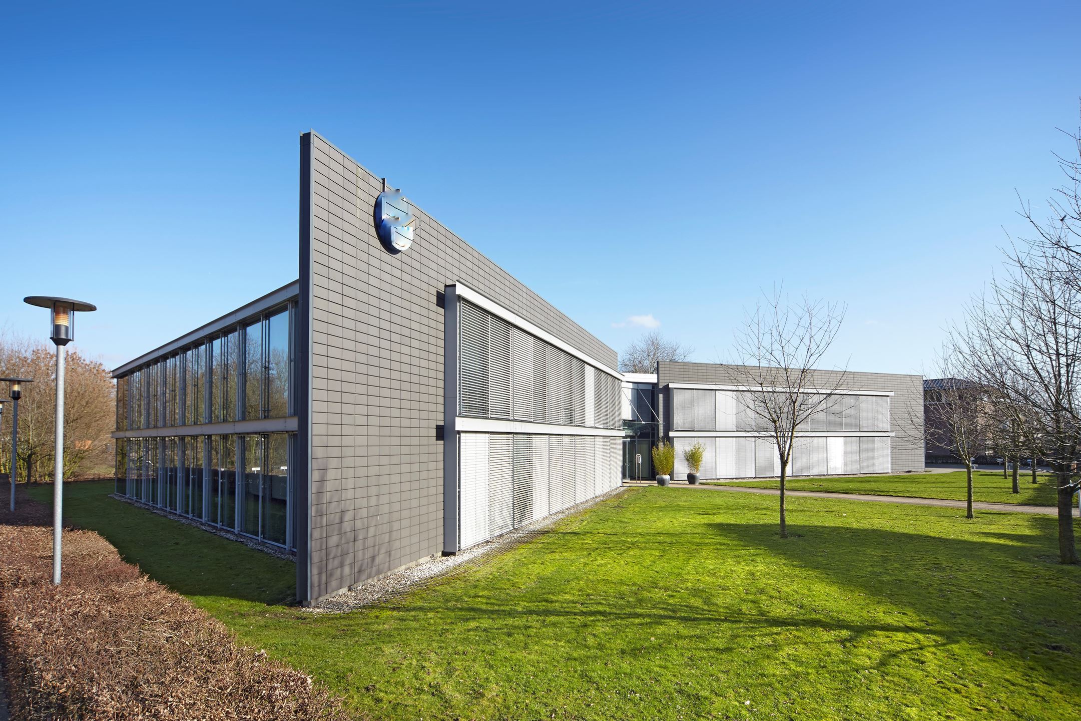 Een kantoorgebouw met twee verdiepingen, grote glazen ramen en strakke architectuur. Gelegen in een goed onderhouden omgeving met grasvelden en jonge bomen. Logo van een bedrijf zichtbaar op de gevel. Rustige en zonnige dag met helderblauwe lucht.
