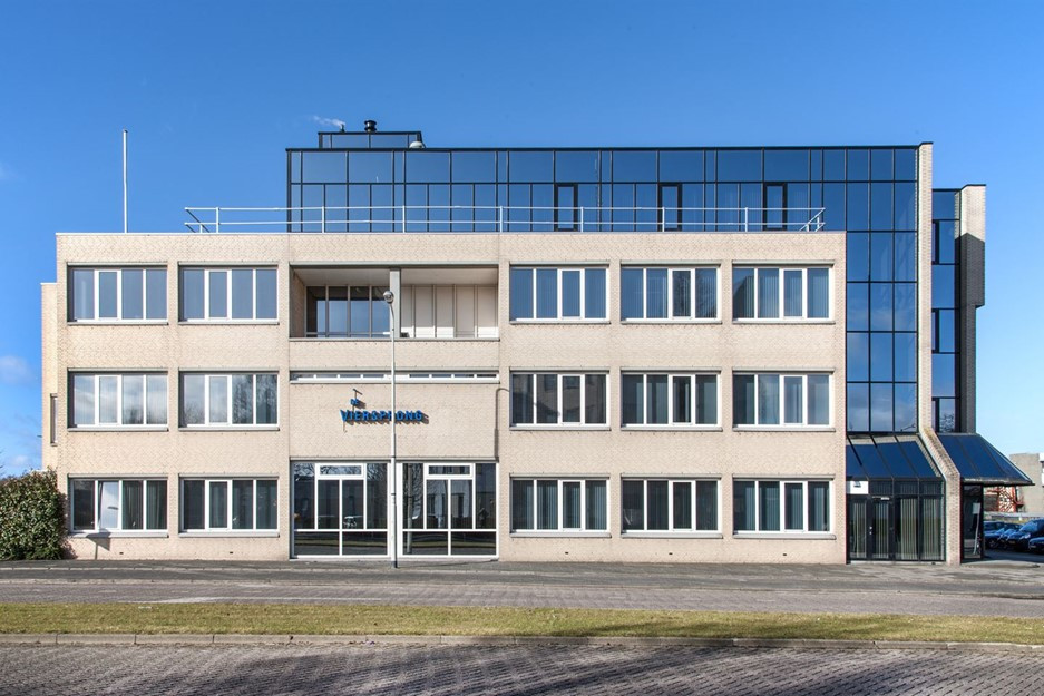 Een commercieel gebouw met drie verdiepingen onder een heldere blauwe hemel. Het gebouw heeft een mix van architectonische materialen en elementen. De eerste twee verdiepingen hebben een lichte beige of gebroken witte gevel, mogelijk van beton of stucwerk.  Het ontwerp van het gebouw is relatief eenvoudig en functioneel, met een plat dak en een rechthoekige vorm. De ramen zijn uniform rechthoekig en gerangschikt in een rasterpatroon, met in totaal vier ramen over en drie ramen naar beneden op elke verdieping. De ramen zijn omlijst in een lichte kleur, wat een visueel contrast creëert met de lichtgekleurde muren.