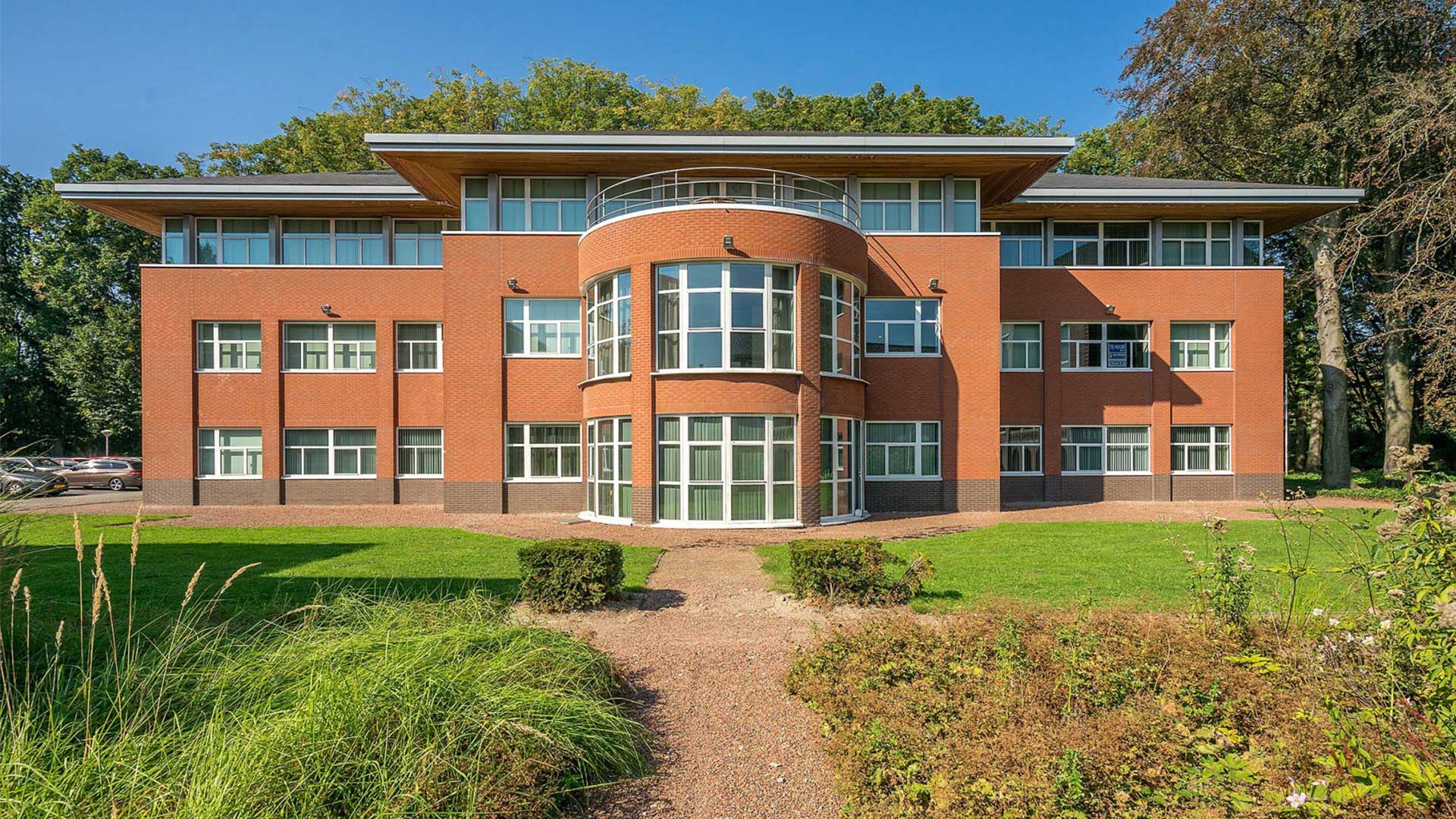 Een kantoorgebouw met drie verdiepingen en een oranje bakstenen gevel. De bovenste etage is voorzien van grote ramen en de kantoortuin is goed verzocht. 