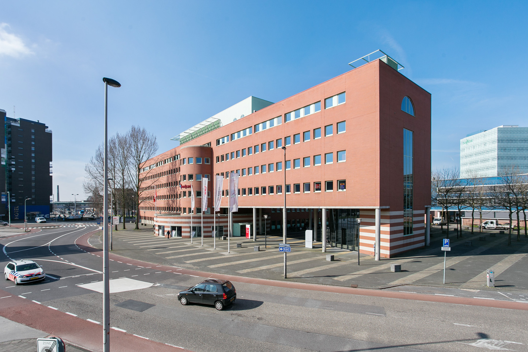 Een gebouw met meerdere verdiepingen. Het gebouw heeft een opvallende baksteenrode kleur en is hoekig van vorm met een gedeeltelijk afgeknotte hoek die een uniek architectonisch detail vormt. Het dak van het gebouw is plat en aan de voorzijde van het gebouw bevinden zich verschillende grote ramen die regelmatig over de gevel zijn verdeeld. Op de begane grond is de muur verdeeld in meerdere lagen van gekleurde stenen rood om wit.  Voor het gebouw staan vier vlaggenmasten. De omgeving van het gebouw is verstedelijkt, met bestrating en een kleine groenstrook met jonge bomen langs de kant van de weg.  Aan de overkant van de weg is nog een groot gebouw zichtbaar, dat een glazen gevel heeft en een moderne uitstraling geeft. De lucht is helder en blauw, wat suggereert dat het een zonnige dag is.  De algemene sfeer van de afbeelding is rustig en geordend, met een focus op moderne architectuur en georganiseerde stedelijke planning.
