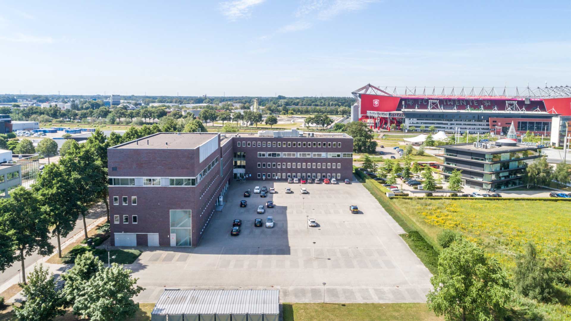 Een uitgestrekte horizon met een bijna strak blauwe lucht. Op de voorgrond zie je een kantoorgebouw en rechts op de achtergrond een voetbalstadion.
