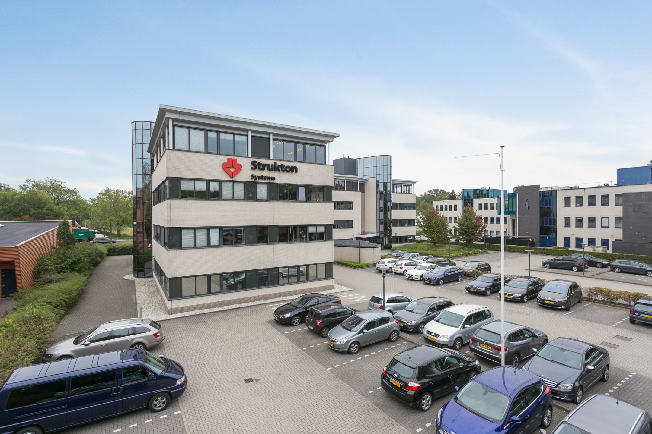 Een commercieel kantoorgebouw van drie verdiepingen met hoekige architectuur, witte gevelpanelen en grote glaspartijen als ramen. Op de gevel staat het 'Strukton Systems' logo. Het gebouw bevindt zich op een heldere dag met enkele wolken in de lucht. Voor het gebouw is een parkeerterrein te zien met verschillende geparkeerde auto's en groene struiken. Een ander modern gebouw is aan de rechterkant zichtbaar. De lucht is blauw met verspreide wolken en een vlaggenmast staat in de buurt. Deze afbeelding straalt een professionele zakelijke omgeving uit tijdens een normale werkdag.