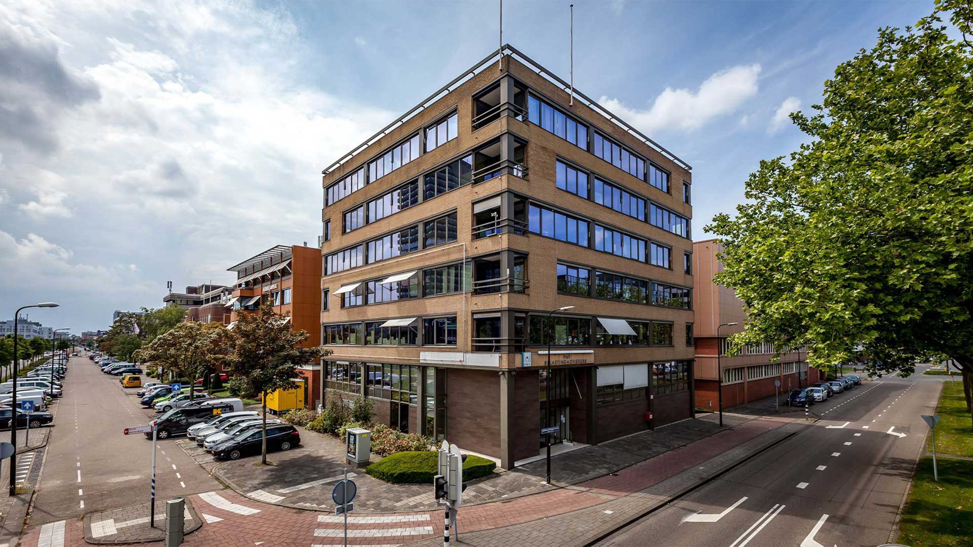 Een stedelijke scène met een modern, meerlaags gebouw met een gevel in beige en bruine tinten en grote ramen. Het gebouw bevindt zich op een hoek en de architectuur omvat enkele overhangende elementen en wat lijkt op een ingang of lobby met een overdekte loopbrug.  De straat naast het gebouw heeft een rij geparkeerde auto's en is omzoomd met bomen. Er is ook een voetgangersoversteekplaats en diverse wegmarkeringen zichtbaar. Aan de rechterkant van de afbeelding staat een grote boom met dicht groen bladerdek dat het zicht op de gebouwen verderop in de straat deels belemmert.  De lucht is deels bewolkt, wat suggereert dat het een dag kan zijn met afwisselend zon en bewolking. De algehele indruk is van een rustig, goed onderhouden stedelijk gebied, mogelijk in een residentiële of gemengd gebruik deel van een stad.