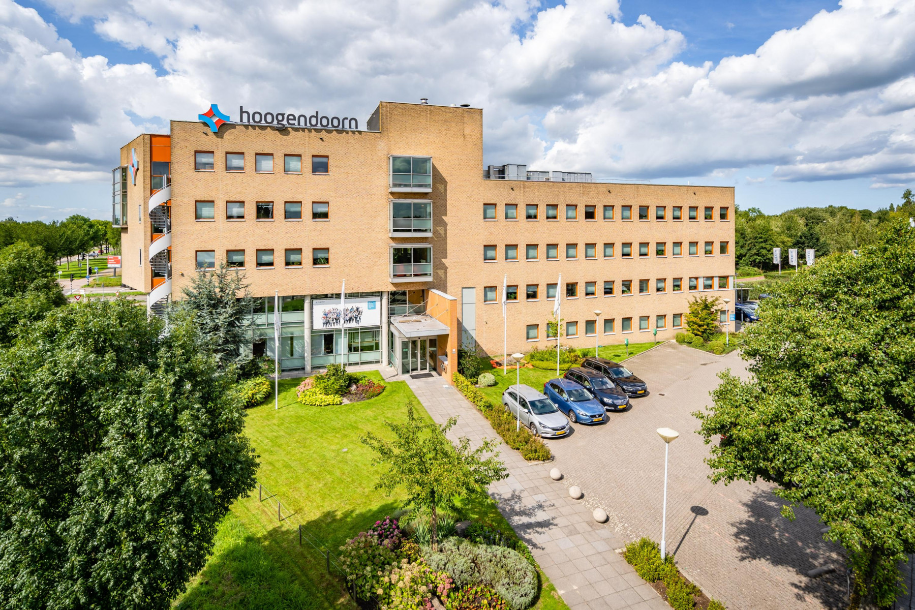 Een modern kantoorgebouw van vier verdiepingen op een zonnige dag met enkele wolken in de lucht. Het gebouw heeft een bruine bakstenen gevel met veel ramen. Op het dak van het gebouw staat in grote letters 'Hoogendoorn'. Voor het gebouw loopt een duidelijk pad dat naar de ingang leidt. Links van het pad ligt een netjes onderhouden groen gazon met enkele bomen en struiken. Aan de rechter voorzijde van het gebouw is een parkeerplaats met verschillende auto's. Het lijkt op een bedrijfsgebouw in een rustige, groene omgeving.