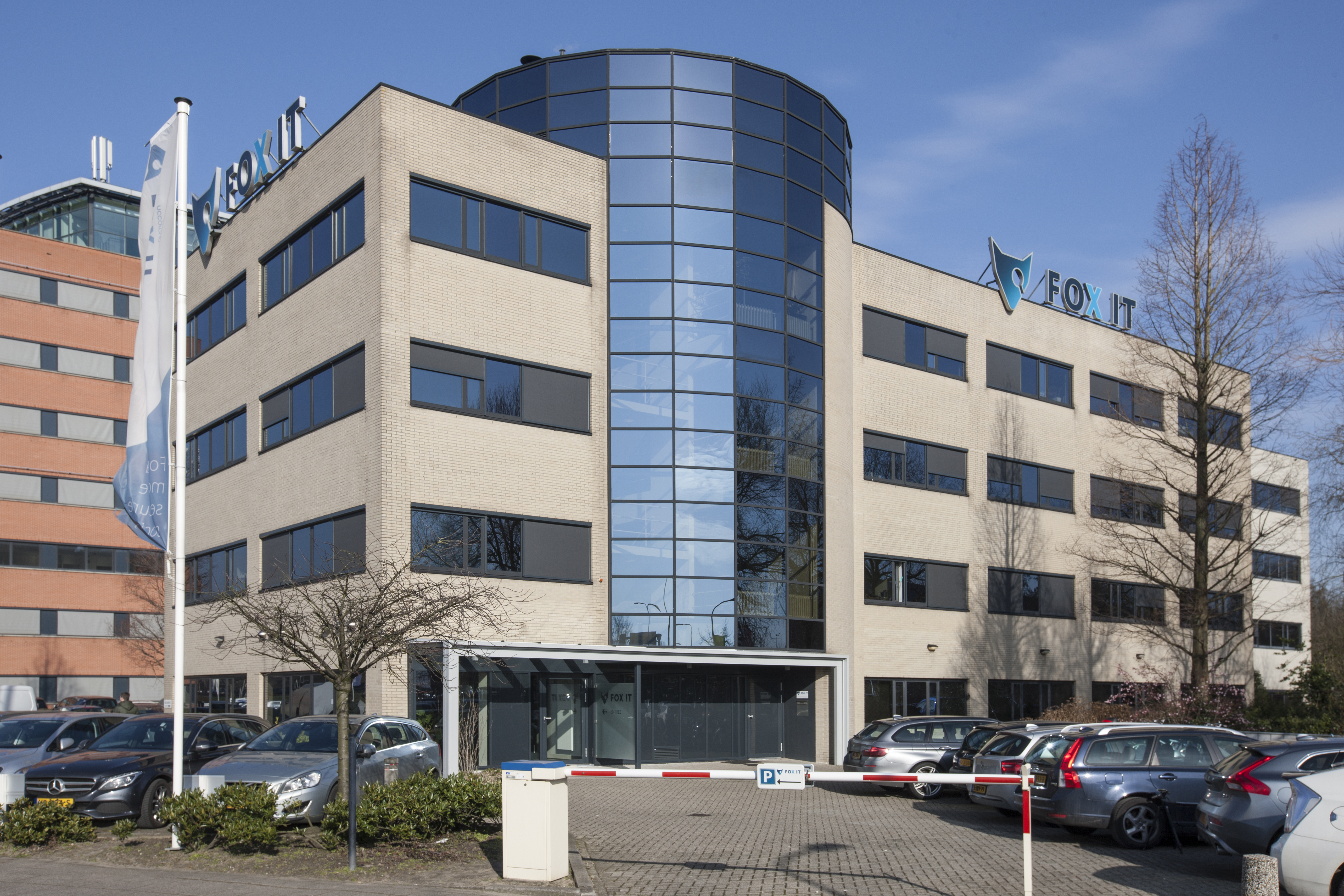 Een kantoor gebouw met grote rechthoekige ramen en een parkeerplaats met slagboom op de voorgrond.