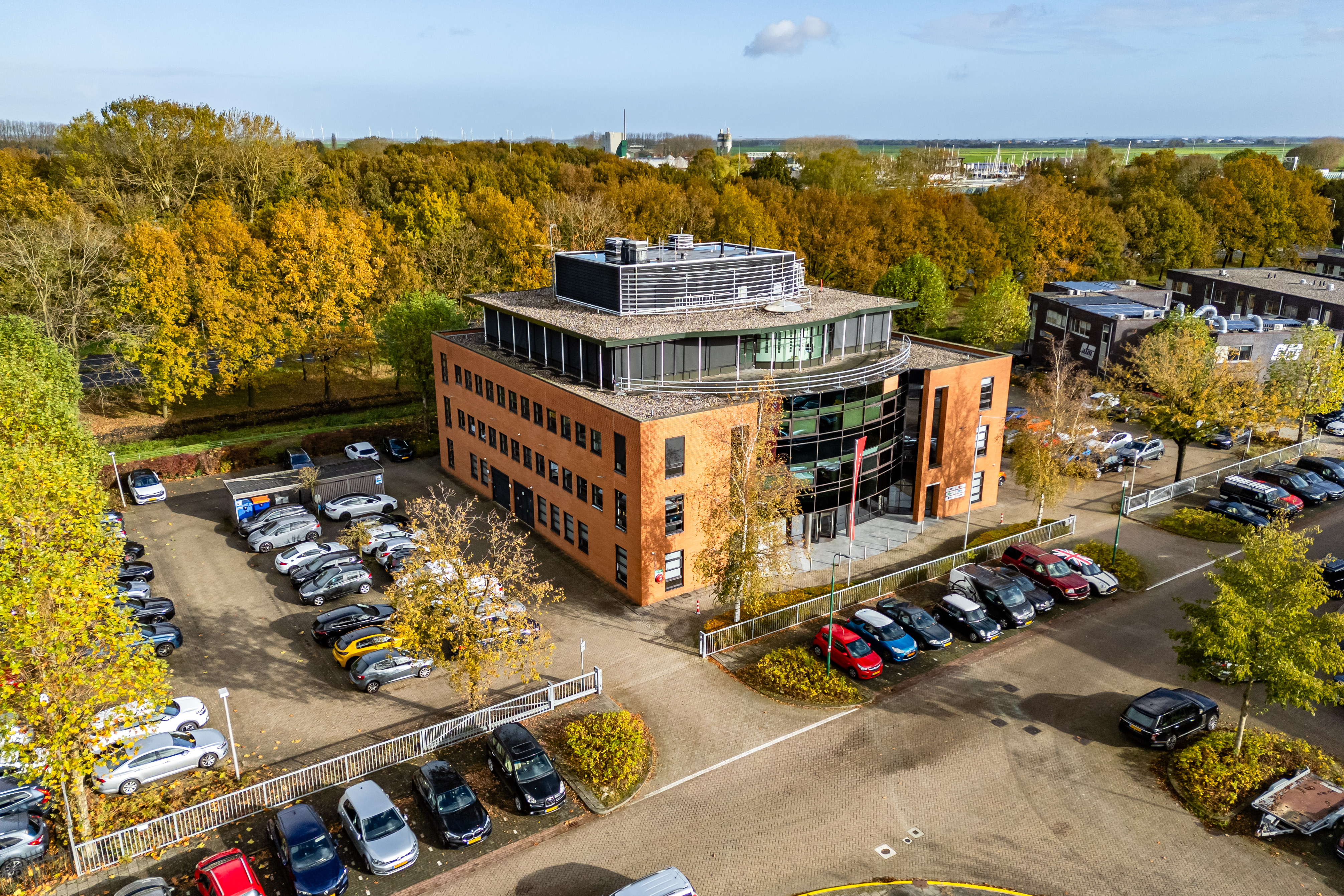 Luchtfoto van kantoorgebouw Baarnsche Dijk 8 in Baarn. Een gebouw met meerdere verdiepingen, parkeervakken rondom en bomen in herfstkleuren.