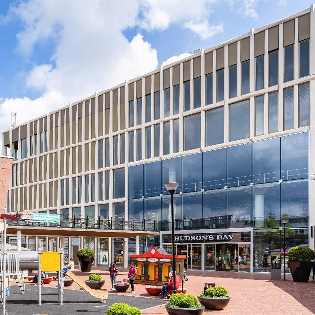 Een modern gebouw met een glazen gevel en een plein ervoor, waar mensen over lopen. Bovenaan het gebouw is de tekst 'HUDSON'S BAY' zichtbaar.
