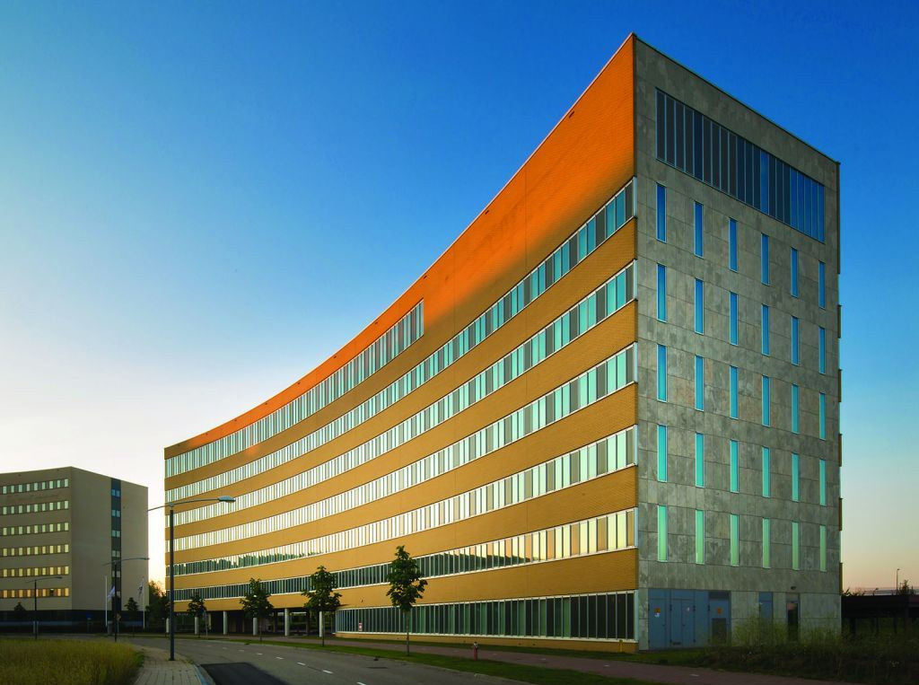 Een gebouwencomplex met meerdere niveaus en secties. De architectuur omvat een mix van rechte en gebogen lijnen, met een opvallende gebogen glazen gevel aan één uiteinde. Het gebouw wordt omringd door groen, wegen en geparkeerde auto’s, wat aangeeft dat het zich in een stedelijk gebied bevindt.