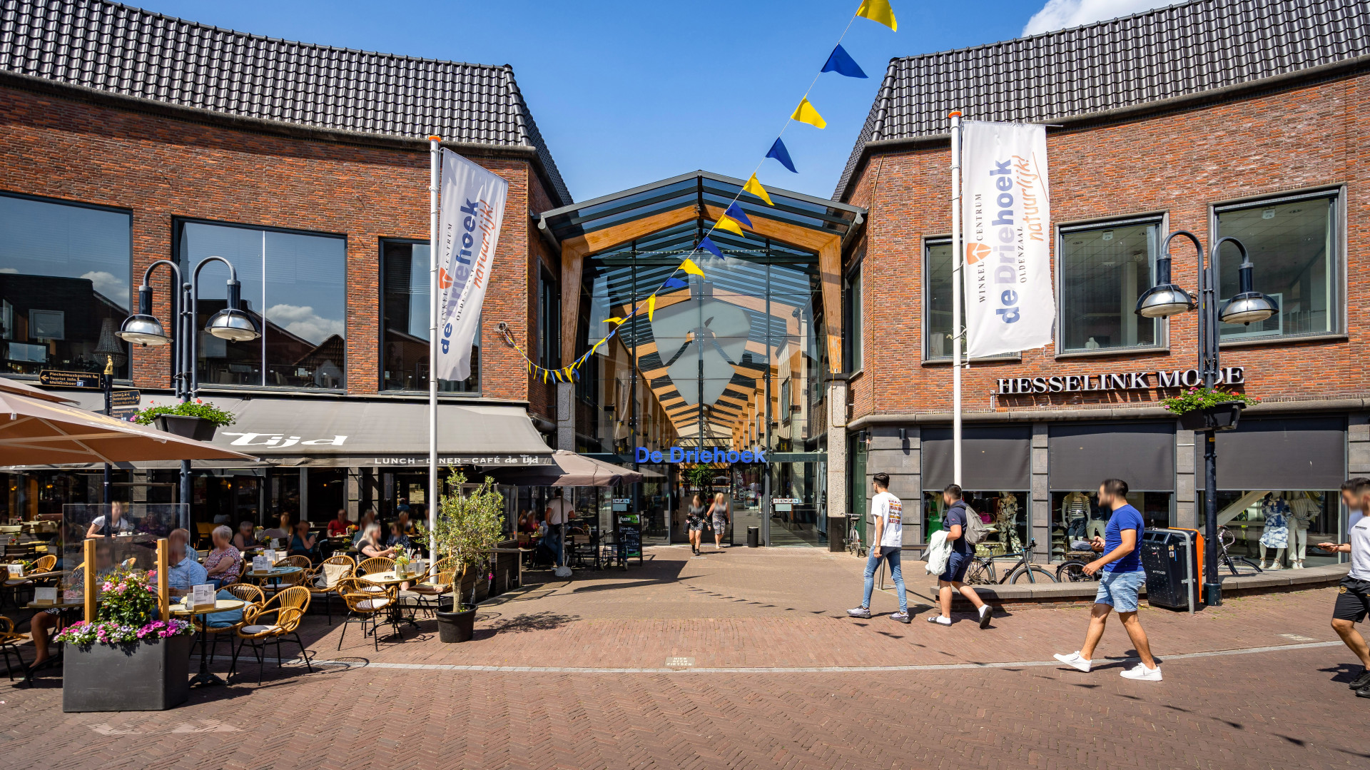 Een levendige voetgangersstraat geflankeerd door bakstenen gebouwen met in het midden een moderne glazen structuur onder een blauwe lucht, met een café met terras en mensen die wandelen, wat wijst op een bruisend stedelijk winkelgebied.