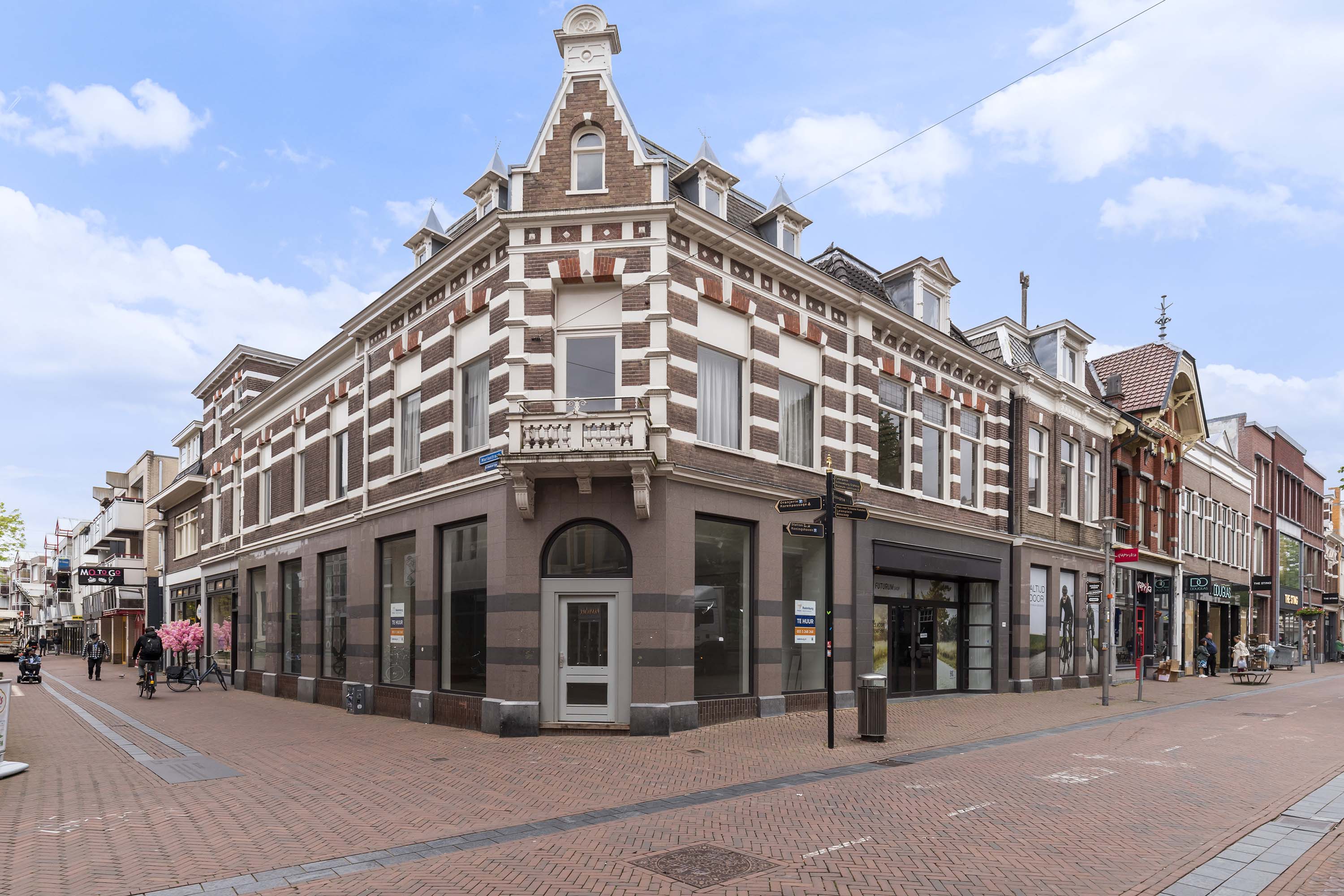 Stedelijke straathoek met prominent hoekgebouw in Europese stijl. Wit gebouw met rode accenten, drie verdiepingen en commerciële ruimtes op de begane grond. Fietsen geparkeerd op de stoep, voetgangers en diverse architecturale stijlen van omliggende gebouwen. Bewolkte lucht, serene stedelijke scène met focus op het hoekgebouw.