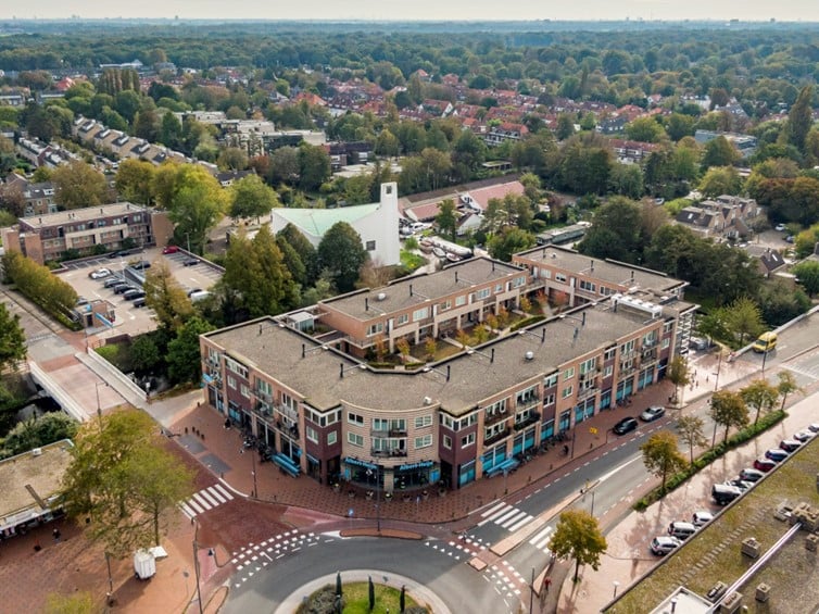 Een luchtfoto van een stedelijk gebied bij daglicht. Het centrale punt van de foto is een drie verdiepingen tellend gebouw met een plat dak, gelegen aan een kruispunt met een kleine rotonde. Het gebouw heeft een onregelmatige vorm met verschillende uitsteeksels en inhammen en lijkt een wooncomplex te zijn, mogelijk met winkels op de begane grond, gezien de aanwezigheid van luifels en uithangborden.  Er zijn diverse auto's geparkeerd langs de straten en enkele auto's rijden op de weg. De straten rondom het centrale gebouw zijn omzoomd met rijen bomen en andere vegetatie.  In de achtergrond zijn veel residentiële huizen te zien, voornamelijk twee verdiepingen hoog, met schuine daken en verschillende kleuren. De huizen zijn omgeven door goed onderhouden tuinen en er zijn meer bomen verspreid over het gebied. De algemene indruk is die van een rustige, goed onderhouden wijk met veel groen. De lucht is overwegend helder met enkele wolken.