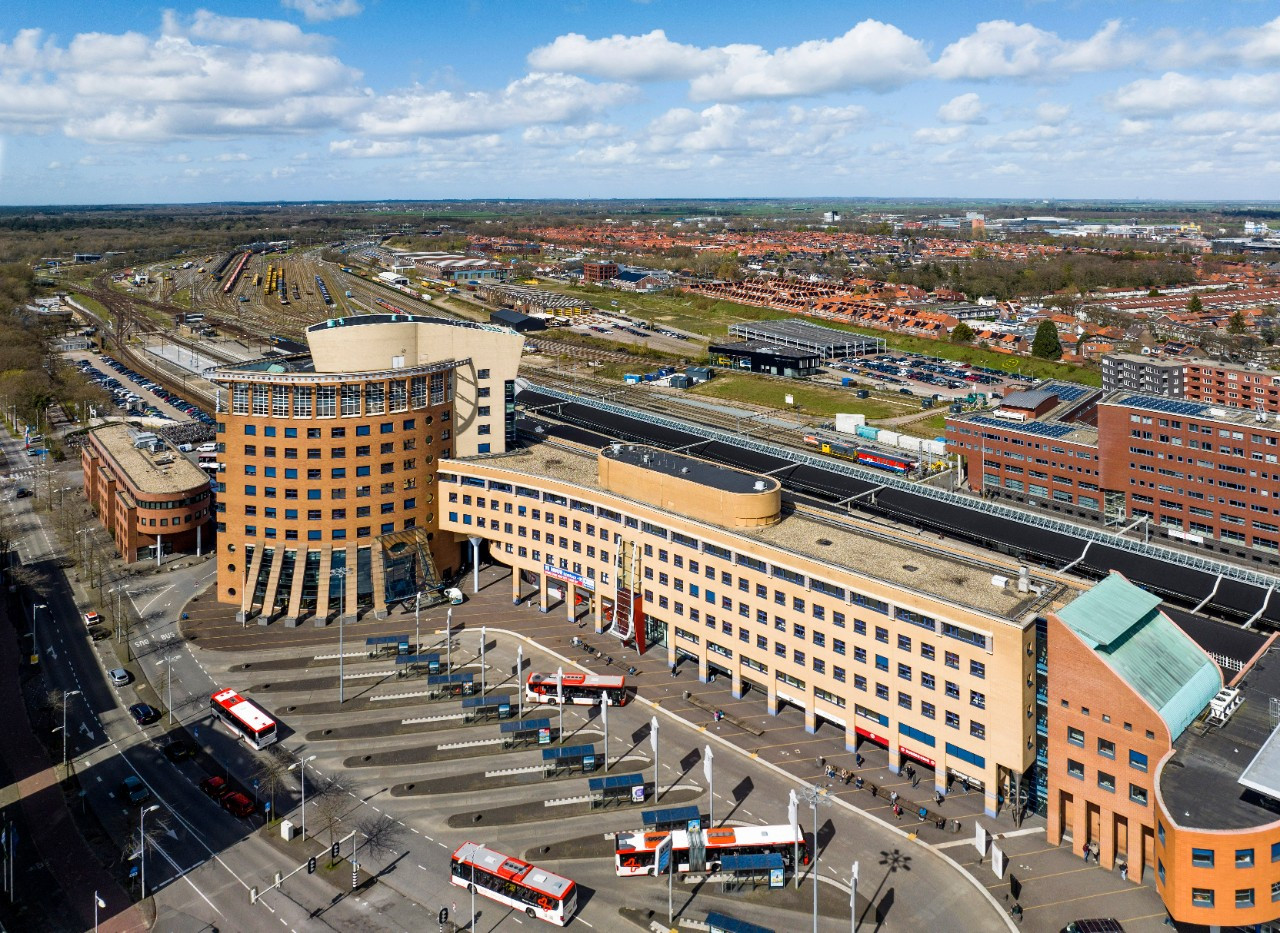 Een luchtfoto, genomen vanuit een drone, van een modern stedelijk gebied tijdens de dag met heldere luchten en enkele wolken. Het middelpunt is een groot, meerdere verdiepingen tellend gebouw met een gebogen architectuur, met een mix van oranje en beige buitenpanelen. Het gebouw heeft een plat dak en talrijke ramen die het zonlicht reflecteren. Het centrale transportknooppunt wordt gekenmerkt door het bus- en treinstation voor en achter het gebouw.  Voor het gebouw is er een ruim plein met geometrische patronen op de grond, bestaande uit lichte en donkergrijze bestratingsstenen. Meerdere trappen verbinden het plein met de ingangen van het gebouw. Er zijn verschillende rode en witte bussen gestopt op aangewezen plekken langs het plein, wat wijst op de aanwezigheid van een busstation.  Aan de linkerkant van het gebouw zijn meerdere spoorwegsporen met bovenleidingapparatuur te zien, wat de locatie van het treinstation aangeeft. Een paar treinen zijn zichtbaar op de sporen.  Over het algemeen toont de afbeelding een goed georganiseerd en levendig transportknooppunt dat fungeert als een belangrijk verbindingspunt voor het openbaar vervoer, waaronder trams, bussen en treinen. Het ontwerp van het gebied suggereert functionaliteit en toegankelijkheid, gericht op de behoeften van forenzen en reizigers.