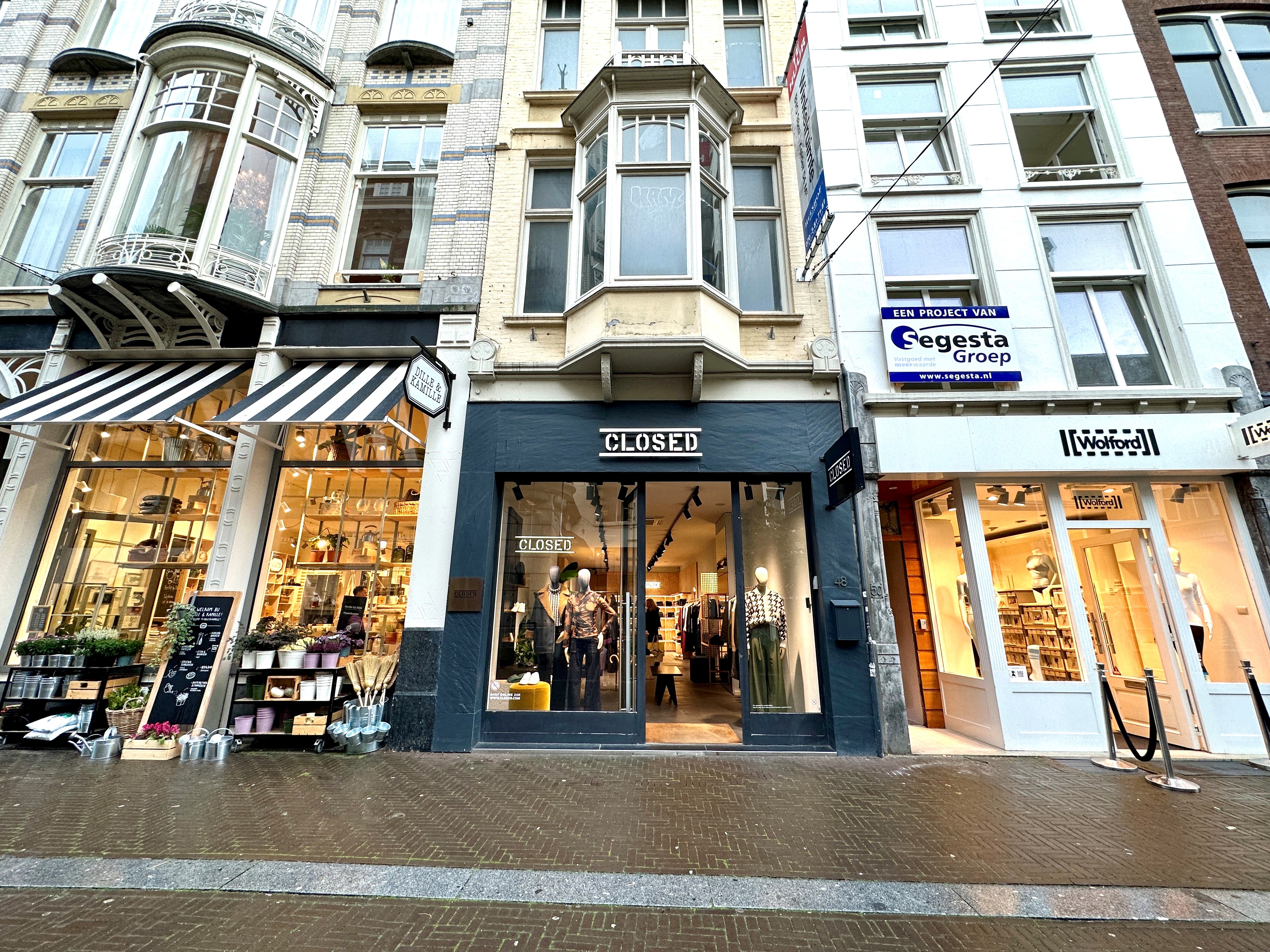 Stedelijk straatbeeld met Europese architectuur en verschillende bedrijfsgevels. Een winkel met 'GESLOTEN' bord, verlichting aan en zichtbare goederen. Naast een bedrijf met zwart-wit gestreept zonnescherm en etalage. Aan de rechterkant een bedrijf met 'Eegesta Groep' bord en 'Noordwijk' aanduiding. Bewolkte lucht, mogelijk vroeg in de ochtend of laat in de middag. Rustige scène zonder mensen of verkeer.
