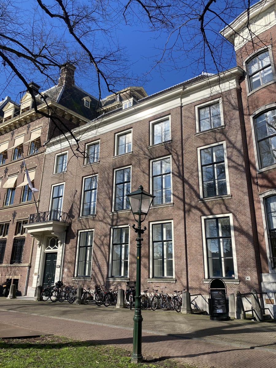 Een traditioneel Europees gebouw met meerdere verdiepingen, bakstenen gevel, boogvormige ramen op de begane grond en witte kozijnen. Fietsen geparkeerd voor de ingang, goed onderhouden grasveld met bladloze bomen, en een zwarte straatlantaarn. Zonnige dag met schaduwen van bomen op het gebouw, wat wijst op ochtend of late middag. Een vredige stedelijke omgeving met een historische sfeer en een mix van geschiedenis en hedendaags leven.
