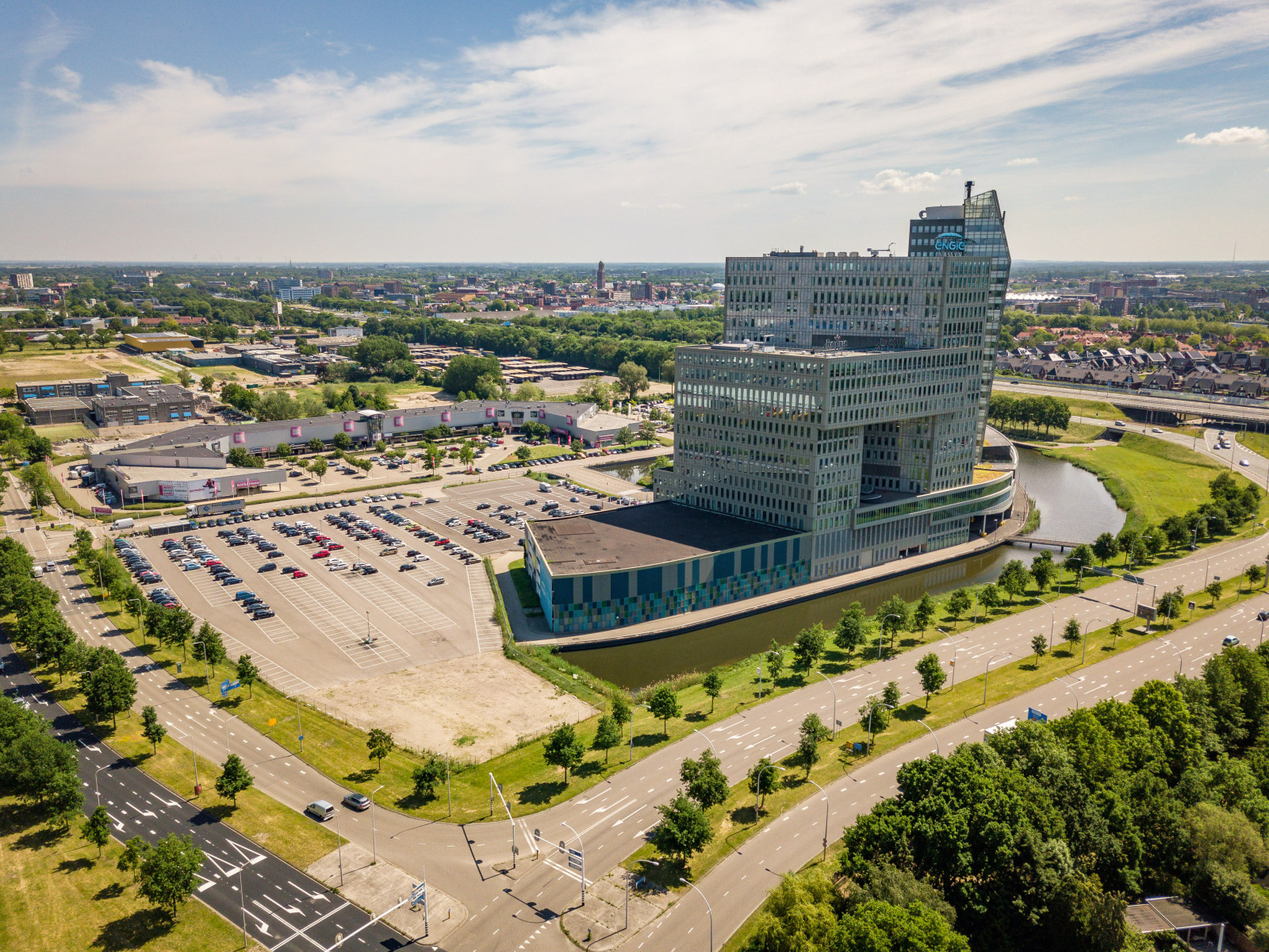 Een luchtfoto die overdag is genomen onder een heldere hemel met enkele verspreide wolken. Het toont een modern stedelijk landschap met een opvallend, uniek ontworpen gebouw als centrale focus.  Het gebouw is meerdere verdiepingen hoog met een onderscheidende, onregelmatige geometrische architectuur, met verschillende uitsteeksels en inkepingen die het een zeer eigentijdse uitstraling geven. De gevel lijkt te bestaan uit glas en metaal, die het zonlicht en de lucht weerspiegelen. Het bevindt zich naast een waterlichaam, wat een sereen milieu creëert en bijdraagt aan de esthetische aantrekkingskracht van de locatie.  Rondom het gebouw is een ruime parkeerplaats gevuld met tal van auto's, wat aangeeft dat het gebouw een plaats van zaken of openbaar belang is die een aanzienlijk aantal bezoekers trekt. De parkeerplaats is goed georganiseerd met gemarkeerde ruimtes en lijkt ongeveer halfvol te zijn.  Verder dan het parkeergebied zijn er groene zones met bomen en gras, die een contrast vormen met de bebouwde omgeving en bijdragen aan de algehele aangenaamheid van de scène.  Samengevat toont de afbeelding een modern en goed gepland stedelijk milieu met een kenmerkend gebouw als middelpunt, omringd door infrastructuur die het gebruik ondersteunt, waaronder een parkeerplaats en aangelegde gebieden, met een breder stedelijk context in de achtergrond.