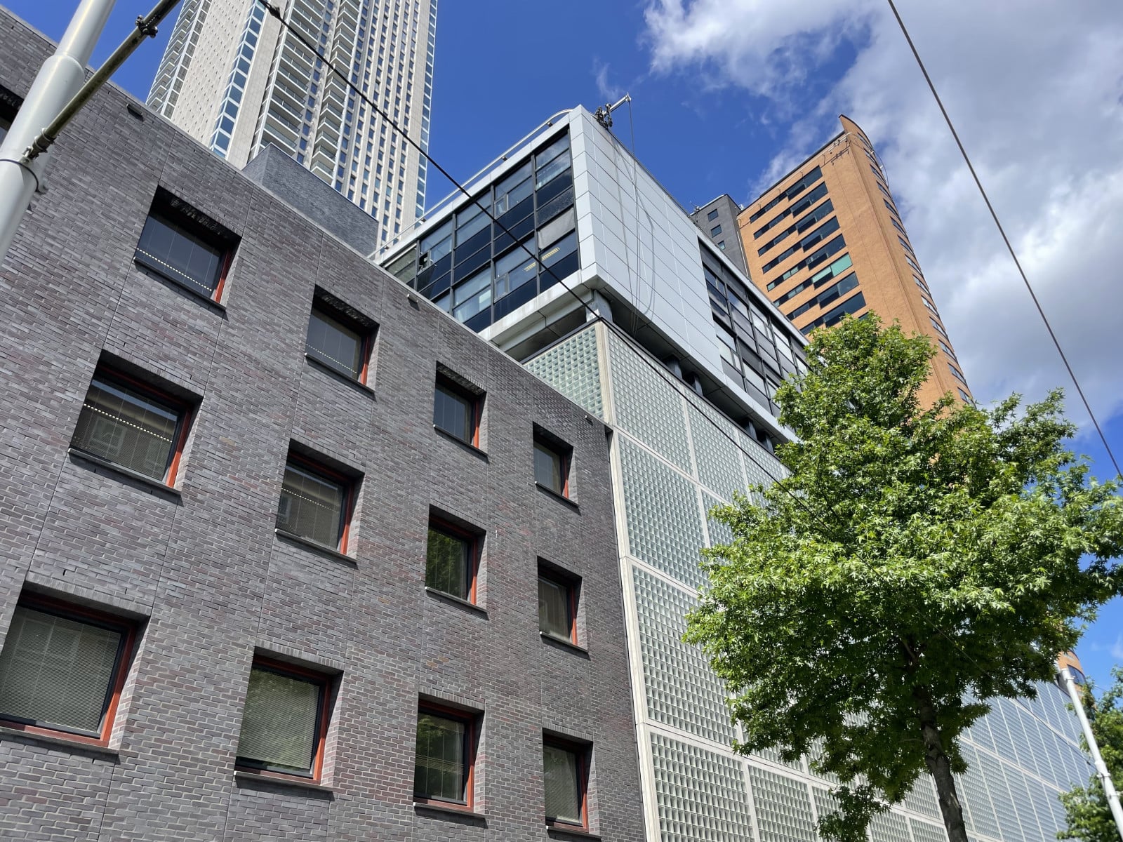 Uitzicht op moderne gebouwen onder een blauwe lucht met verspreide wolken. Op de voorgrond is een gebouw te zien met een grijze gevel en meerdere vierkante ramen. Daarachter staan hogere gebouwen met reflecterende glazen oppervlakken en zichtbare structurele elementen, waarvan er één een oranje-bruine kleur heeft. Deze stedelijke scène is interessant vanwege het contrast tussen de architectonische stijlen en materialen, evenals de opname van natuurlijke elementen in een stadslandschap.