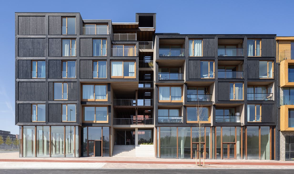 Modern appartementencomplex met symmetrisch ontwerp, voorzien van donkere en lichte houten bekleding, grote glazen ramen en balkons met glazen balustrades tegen een heldere blauwe lucht.