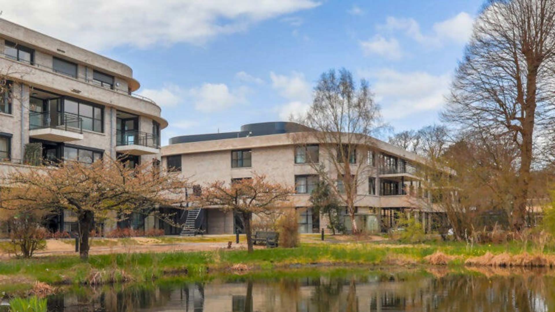 Een modern gebouwencomplex met een organische, ronde architectuur, omgeven door bomen en groen. De verschillende gebouwen met grote ramen staan aan een meer of vijver, waarin ze worden weerspiegeld. De omgeving oogt rustig en natuurlijk, met paden en bankjes voor voetgangers.