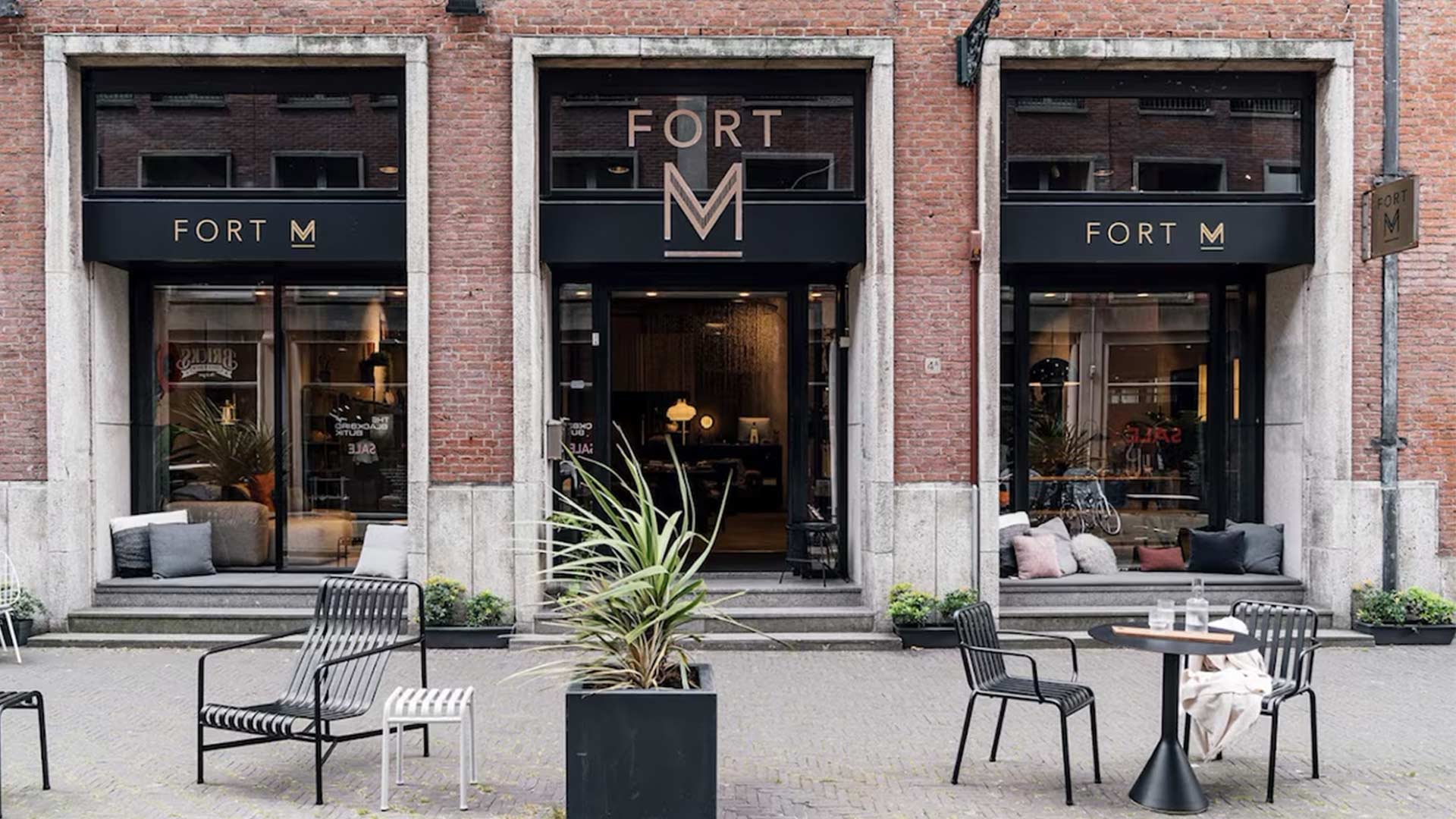 Beeld van een modern bakstenen gebouw met drie ingangen, elk met de naam 'FORT M' erboven. Voor het gebouw staan tafels en stoelen, wat erop wijst dat het een café of restaurant is. Er staat ook een plant in een pot bij de ingang. De omgeving lijkt een stedelijke omgeving te zijn, met andere gebouwen op de achtergrond.