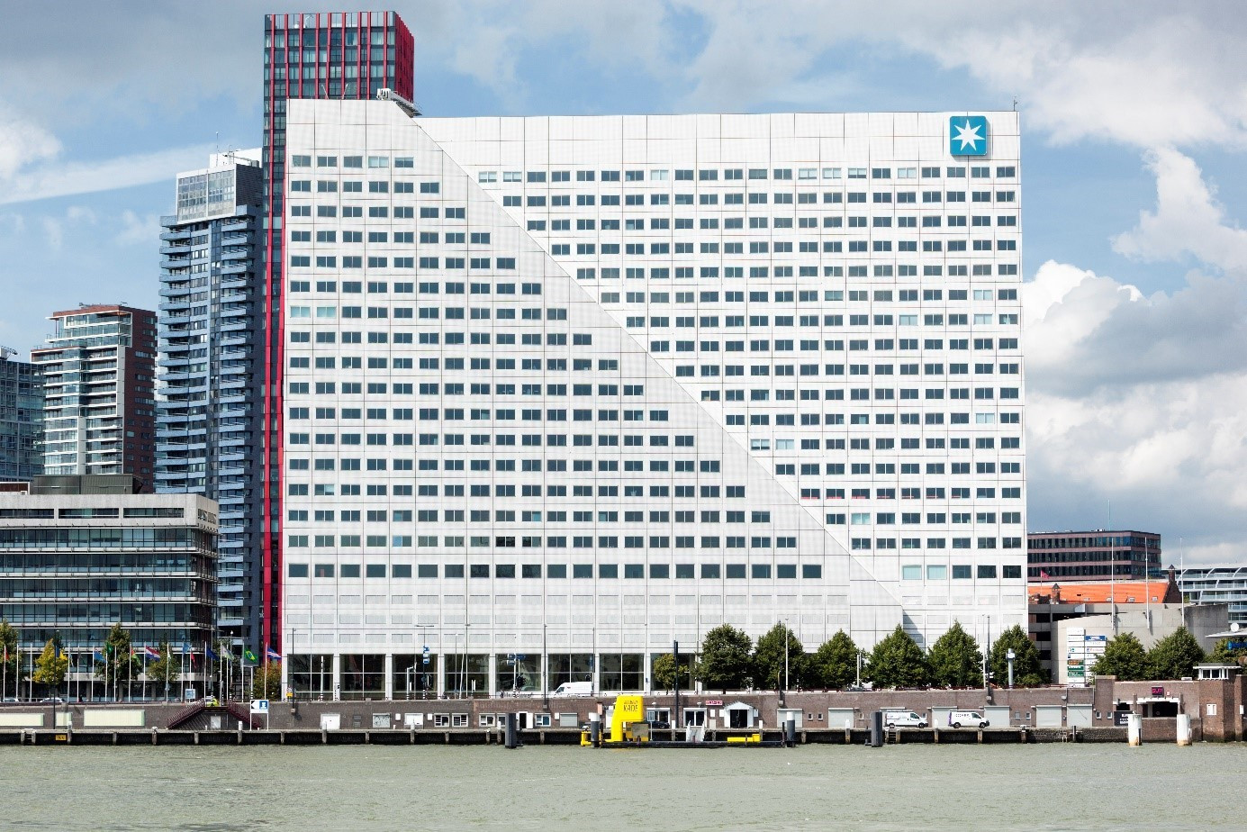 Een modern gebouw met een opvallende architectuur aan de waterkant. Het gebouw heeft een witte gevel met een groot aantal ramen die een zigzagpatroon volgen, wat resulteert in een opvallende diagonale lijn die het gebouw in tweeën lijkt te delen. Bovenaan het gebouw, rechts van het midden, is een groot blauw logo met een witte heptagram, wat duidt op de aanwezigheid van een bedrijf of organisatie met dat symbool als merk.