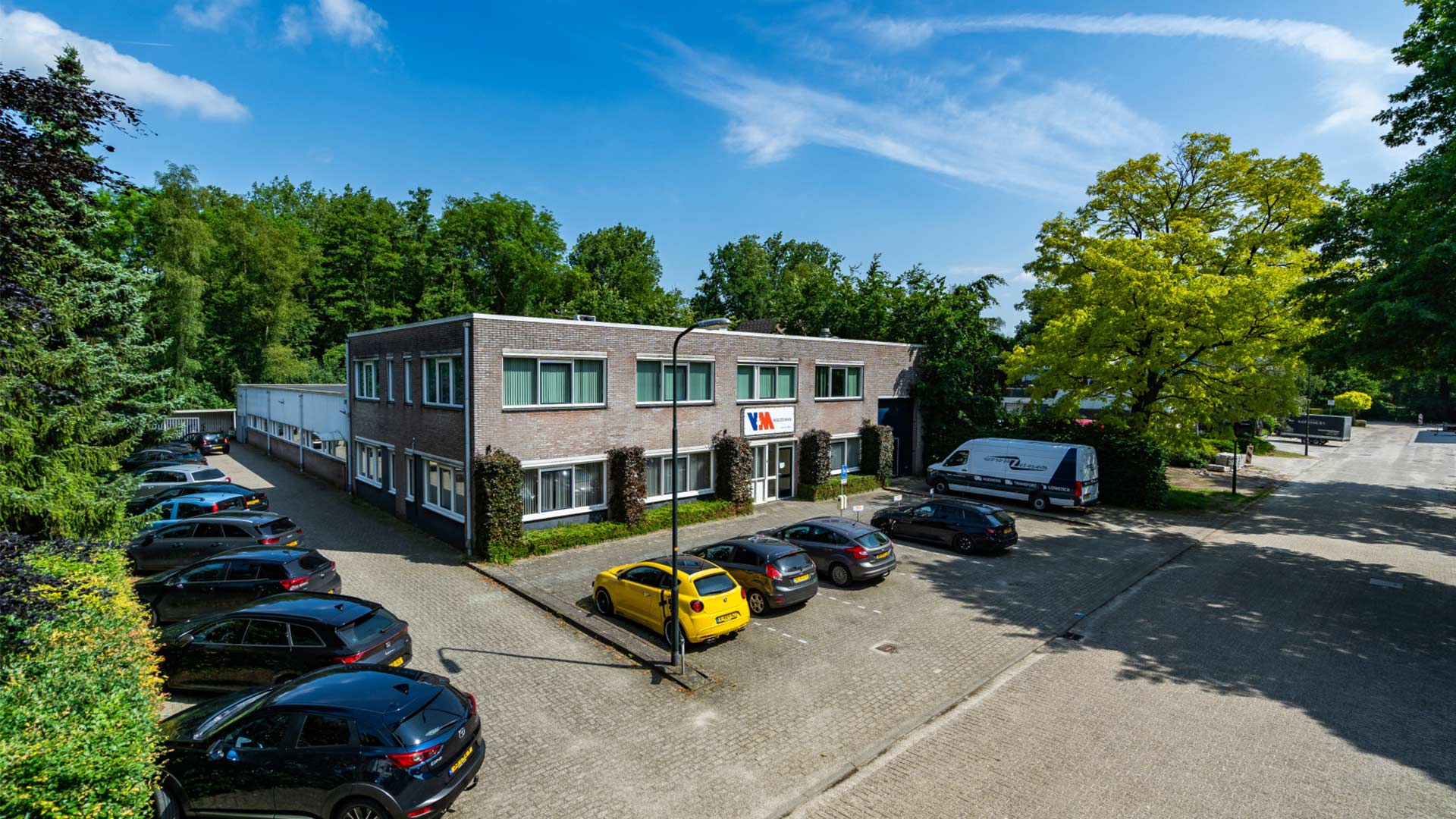 Een modern kantoorgebouw met een parkeerplaats voor het pand, omgeven door groen. Verschillende auto's, waaronder een gele, staan op de parkeerplaats.